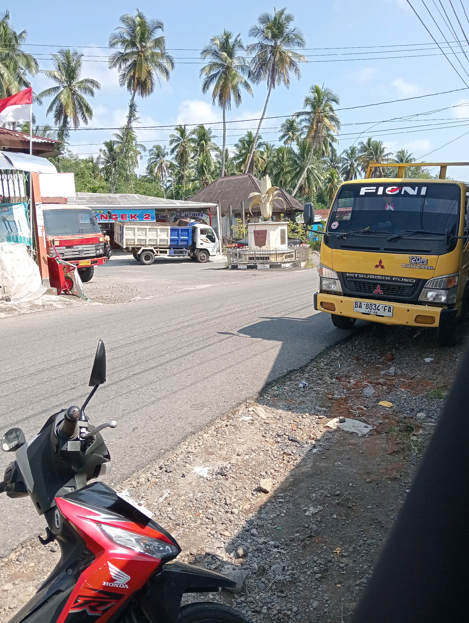 The image captures a street scene in Campago, Indonesia, under a clear blue sky with scattered clouds. The foreground features the front of a red and black Honda motorcycle, partially obscuring a gravelly roadside. Beyond the motorcycle, a paved road runs horizontally, with a yellow Mitsubishi Fuso truck parked on the right side. The truck has the word "FIONL" written on its windshield and license plate "BA 8834 FA". On the left side of the road, a red truck with a blue bed and a white cab is visible, parked near a building with a sign that reads "KAN NGEK 2". Several palm trees are scattered throughout the scene, with a cluster of them in the background behind the buildings. A small monument or statue is also visible behind the red truck, adorned with a gold-colored sculpture. Electrical wires run across the upper portion of the image, suspended between poles. The ground on the right side of the road is covered in gravel and some debris.