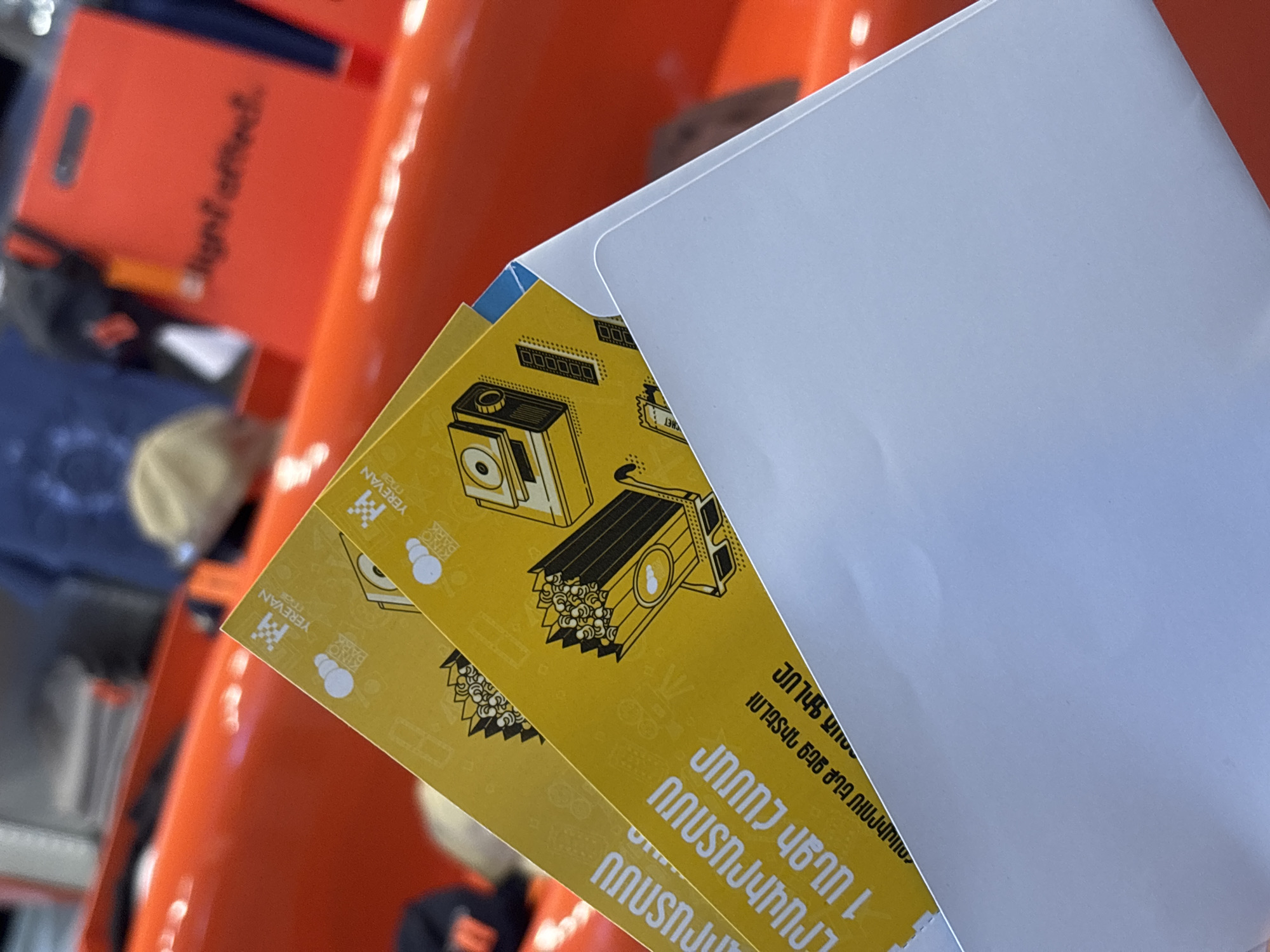 The image shows two yellow tickets with black and white illustrations of vintage-style music equipment in a white envelope. The tickets have Armenian text that is partially visible. The text includes "YEREVAN MALL" and "KINO PARK," suggesting that the tickets might be for a film at a cinema complex in Yerevan Mall. The background appears to be blurred, but it is possibly inside or near a retail or commercial space due to the orange shelving and objects visible in the upper left corner of the image. The context of "Gyumri, Armenia" isn't evident from the image alone.