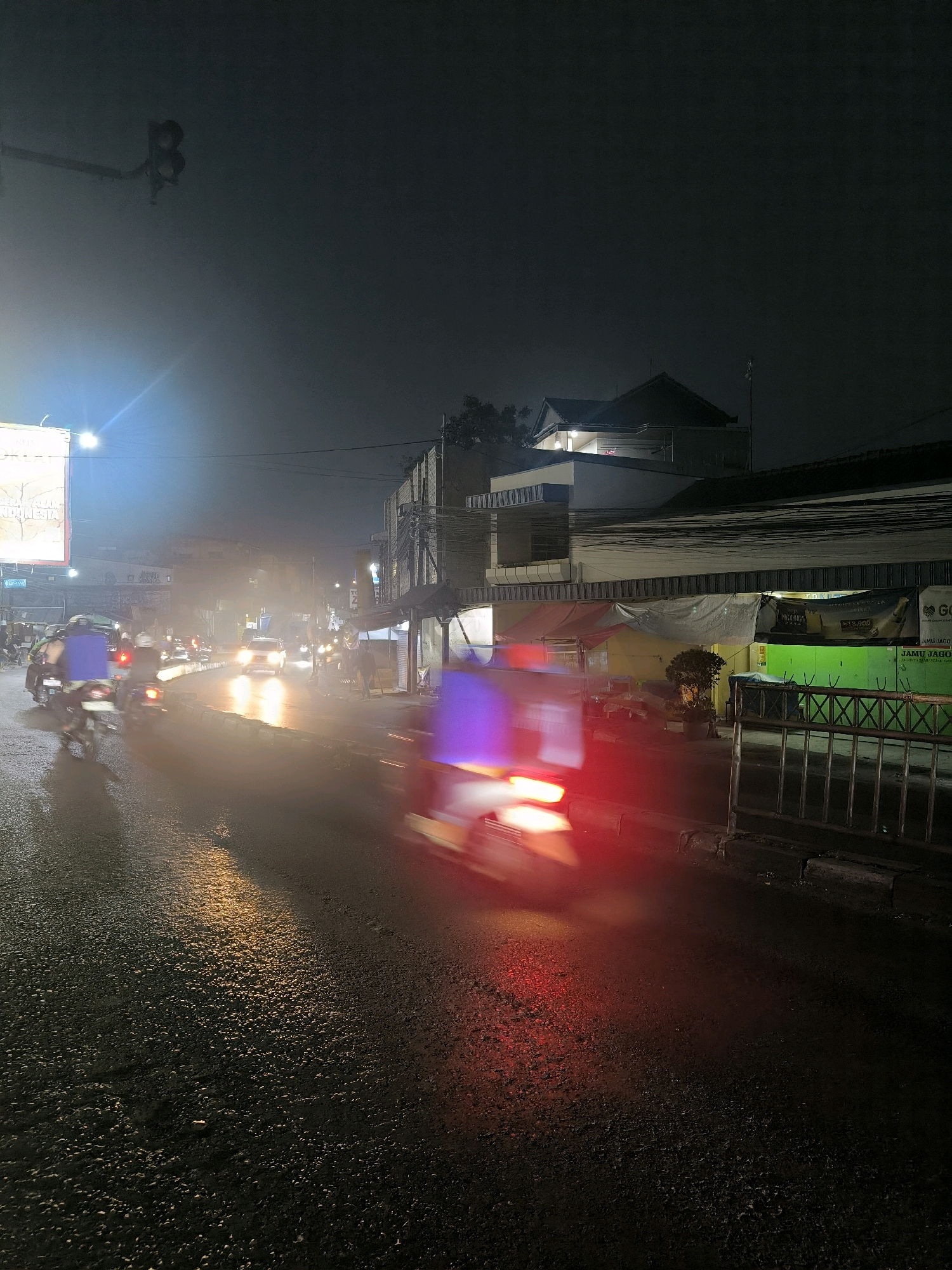 A nighttime, slightly hazy street scene in Bandung, Indonesia, features a two-lane road with wet asphalt reflecting streetlights and vehicle lights. Several motorcycles, some with riders and cargo boxes, are in motion, creating motion blur. One motorcycle in the foreground has a purple illuminated cargo box. A white car is visible further down the road. Buildings line the street, with some showing illuminated windows and signage. A bright, rectangular advertisement is visible on the left. The overall atmosphere is that of a busy urban street at night.