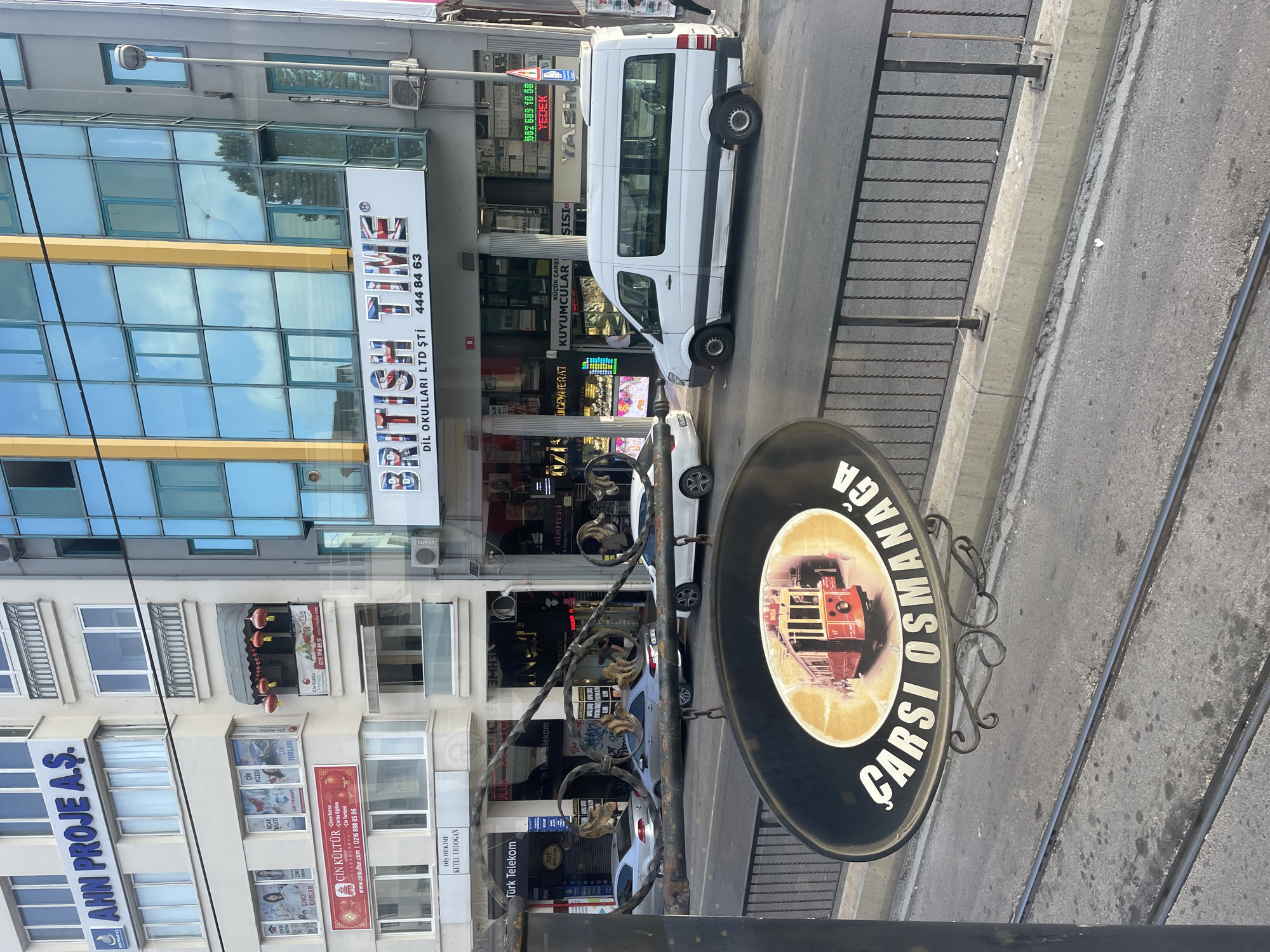 The image shows a street scene in Kadıköy, Turkey, specifically in the vicinity of the Çarşı building. In the foreground, a black oval sign hangs, depicting a vintage red tram with the number 47 and the text "ÇARŞI OSMANAGA" written around it. Behind the sign, tram tracks are visible on the asphalt road.
The mid-ground features a street with several vehicles, including a white van and two silver cars. A black metal railing separates the sidewalk from the street.
The background displays a row of buildings. One building has a prominent sign that reads "BRITISH TIME DİL OKULLARI LTD ŞTİ" with the phone number 444 84 63. Another building is adorned with blue glass windows and a yellow trim, with advertisements for various businesses visible below. To the left, a multi-story building features signage for "AHN PROJE A.Ş." and advertisements related to "ÇİN KÜLTÜR" (Chinese Culture). The environment appears to be an urban setting with shops and commercial establishments lining the street. The overall lighting suggests it is daytime, likely late morning or afternoon.