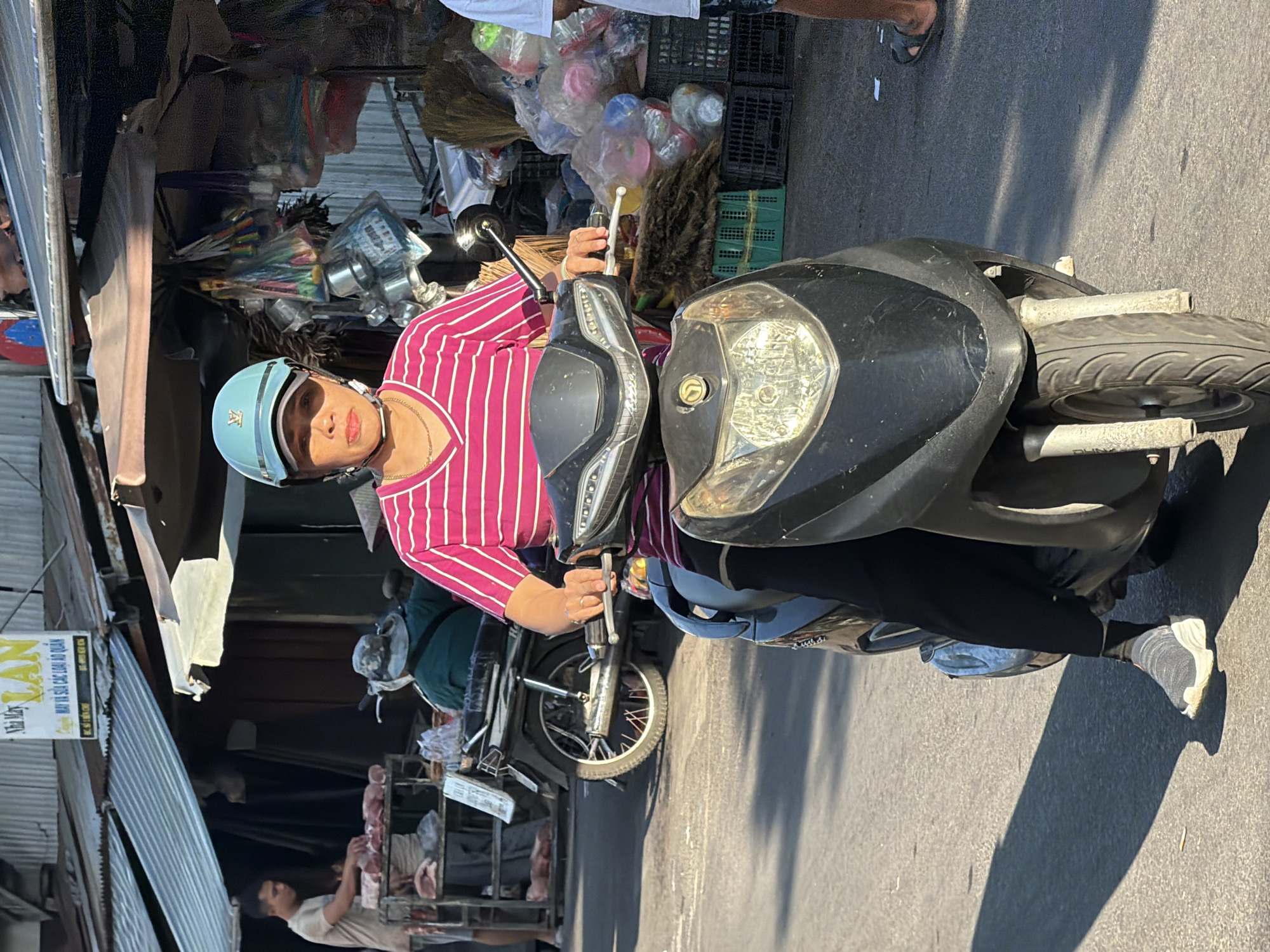 A woman wearing a pink and white striped shirt and black pants is riding a black scooter in what appears to be an outdoor market in Nha Trang, Vietnam. She is wearing a light blue helmet and a gold necklace. The scooter's handlebars are visible, as is the front of the vehicle. Behind her, a man in a tan shirt is seen working at a stall with cuts of meat. Other market stalls with various goods are visible in the background, as are other people and motorbikes. The scene is illuminated by sunlight, casting shadows on the asphalt road.