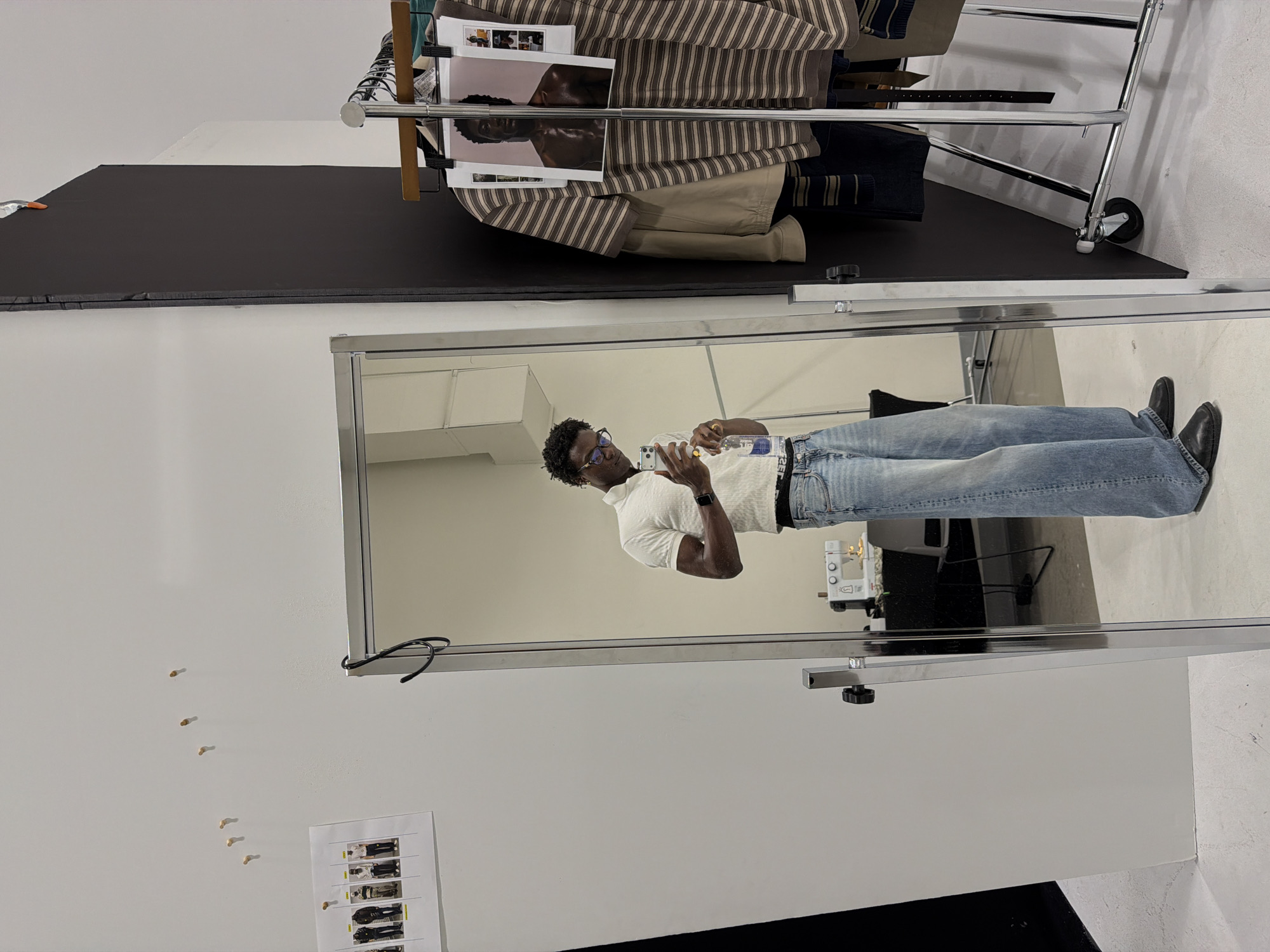 A young Black man is taking a mirror selfie in what appears to be a fitting room or a studio. He is wearing a white short-sleeved polo shirt, blue wide-leg jeans, and black loafers. He is holding a clear plastic bottle of water in his right hand and a smartphone in his left hand. His left arm is bent, and he is looking at his reflection. In the background, there is a sewing machine on a table, a white wall with several pins and a sheet of paper with images of people, and a clothing rack with several garments. The man's reflection is clearly visible in a large, full-length mirror. The overall scene suggests a casual, behind-the-scenes moment, possibly before or after a photoshoot or an event. The city of New Albany, United States of America, is mentioned as the location, but there are no specific visual cues in the image to confirm this.