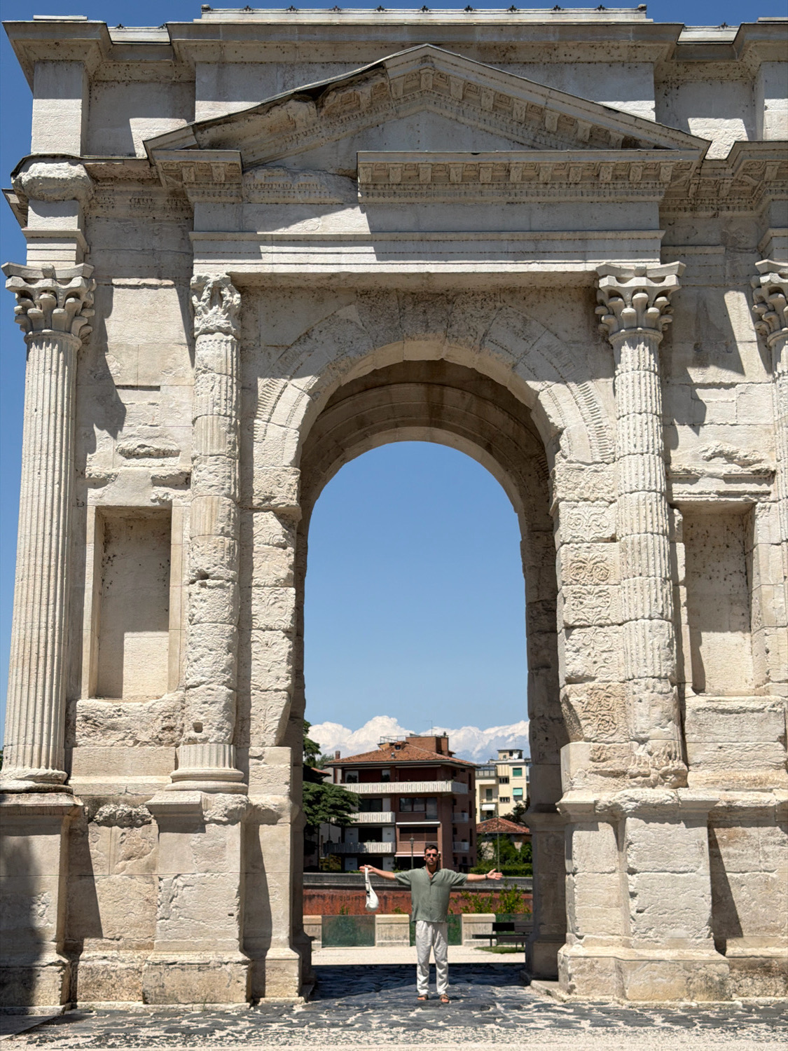 The image shows a man standing in front of the Arch of the Gavi in Verona, Italy. The arch is made of stone and is decorated with carvings. The man is wearing a green shirt and white pants and is holding a white bag. He has his arms outstretched. In the background, there are buildings and mountains. The sky is blue and clear.