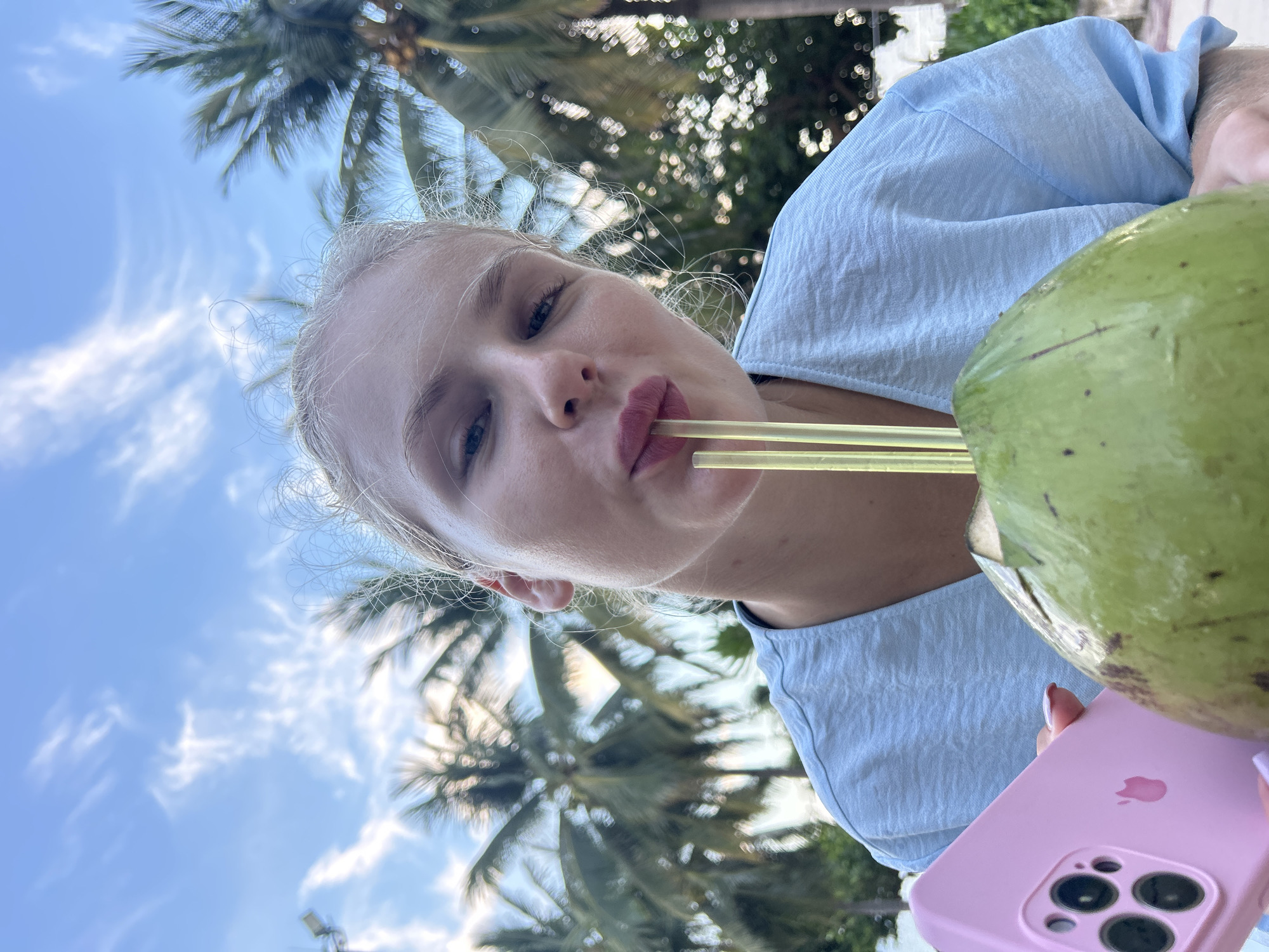 A young Caucasian woman with blonde hair pulled back is drinking from a green coconut with three yellow straws. She is wearing a light blue t-shirt and holding a pink iPhone in her left hand. The background consists of palm trees and a cloudy blue sky, suggesting an outdoor tropical setting. The image appears to have been taken in Nha Trang Ward, Vietnam.
