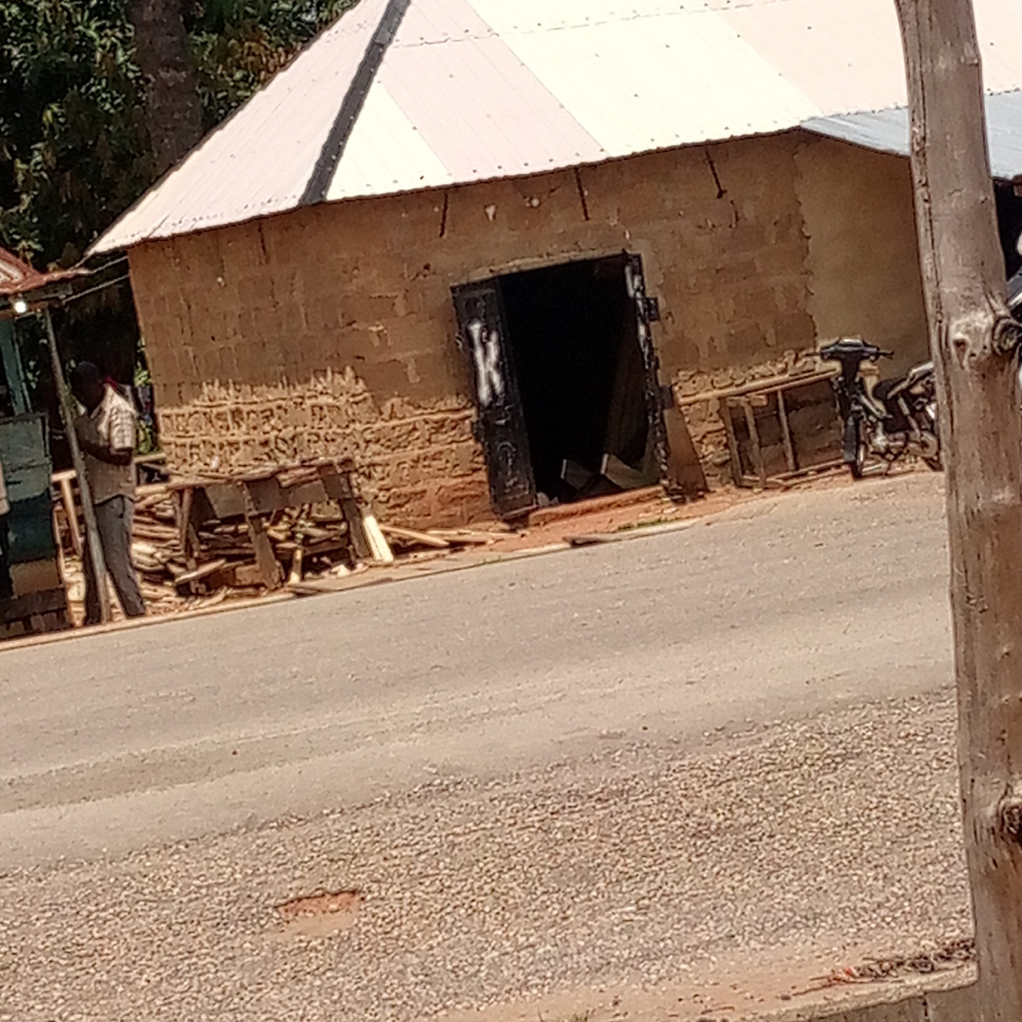 The image presents a street scene in Mararaba, Nigeria. A rectangular, single-story building with a light-colored corrugated metal roof dominates the background. The building's exterior is constructed from rough, earthen blocks. A dark doorway is partially visible in the building's front, marked with some white painted letters, including the letter "K." To the left of the building, a dark-skinned man wearing a light-colored shirt and dark pants stands near some wooden planks and structures. A motorcycle is parked on the right side of the building, partially obscured by what appears to be a thick tree trunk in the foreground. The road in front of the building is made of asphalt and shows signs of wear. A gravel shoulder is visible at the edge of the road in the foreground. The setting appears to be a commercial area, possibly a small workshop. The lighting suggests it is daytime.