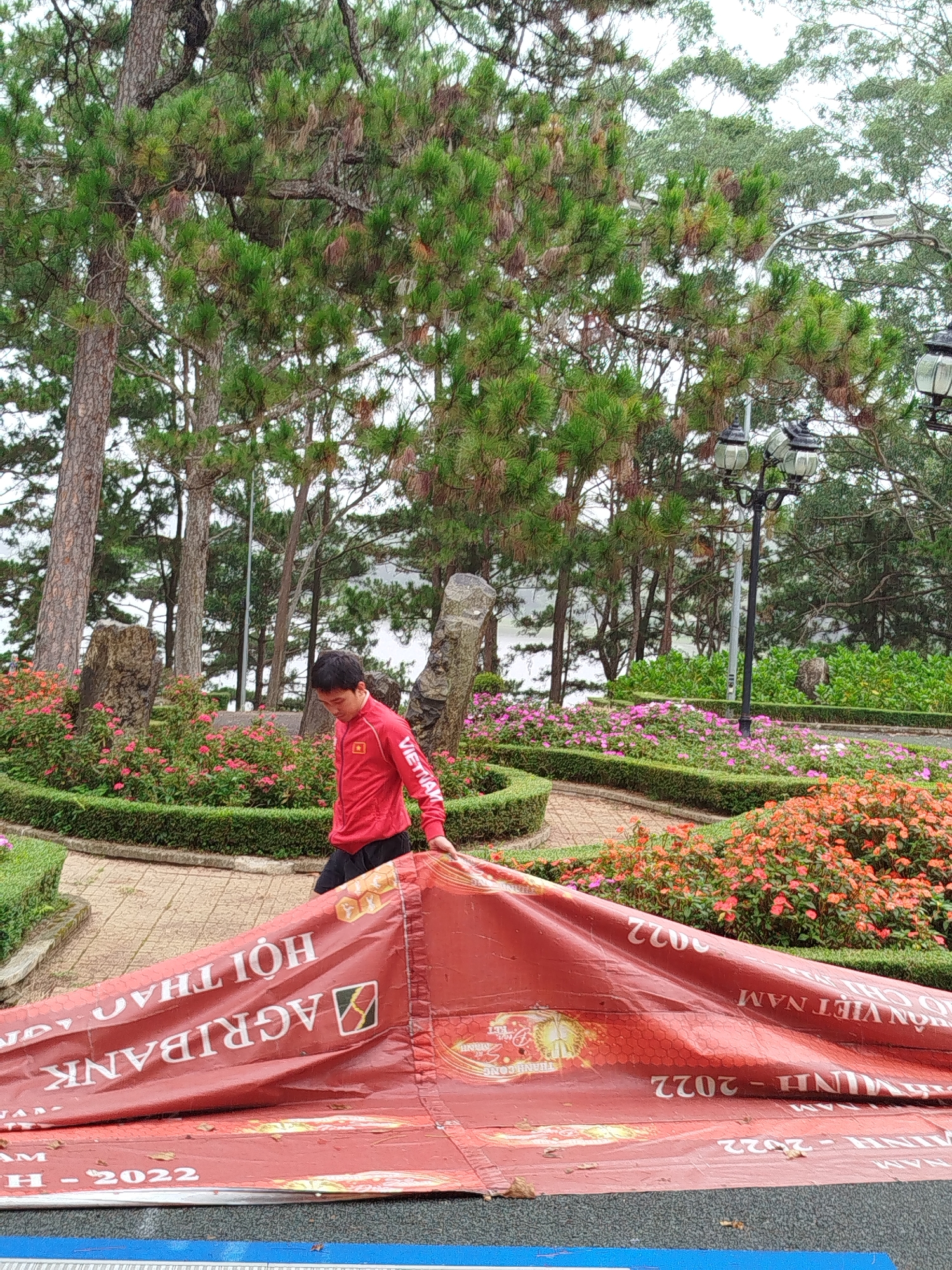 A young man in a red jacket with "VIETNAM" written on the sleeve is in the process of unfolding a large red banner in a park in Da Lat, Vietnam. The banner has white text and logos on it, including "AGRIBANK". The park is characterized by lush green pine trees, manicured hedges, and flower beds filled with pink and orange blossoms. The ground is paved with brick-like tiles, and there are decorative stone sculptures visible. In the background, a lake or body of water can be seen through the trees, and several ornate lampposts are mounted on poles. The sky is overcast, suggesting a cloudy day.
