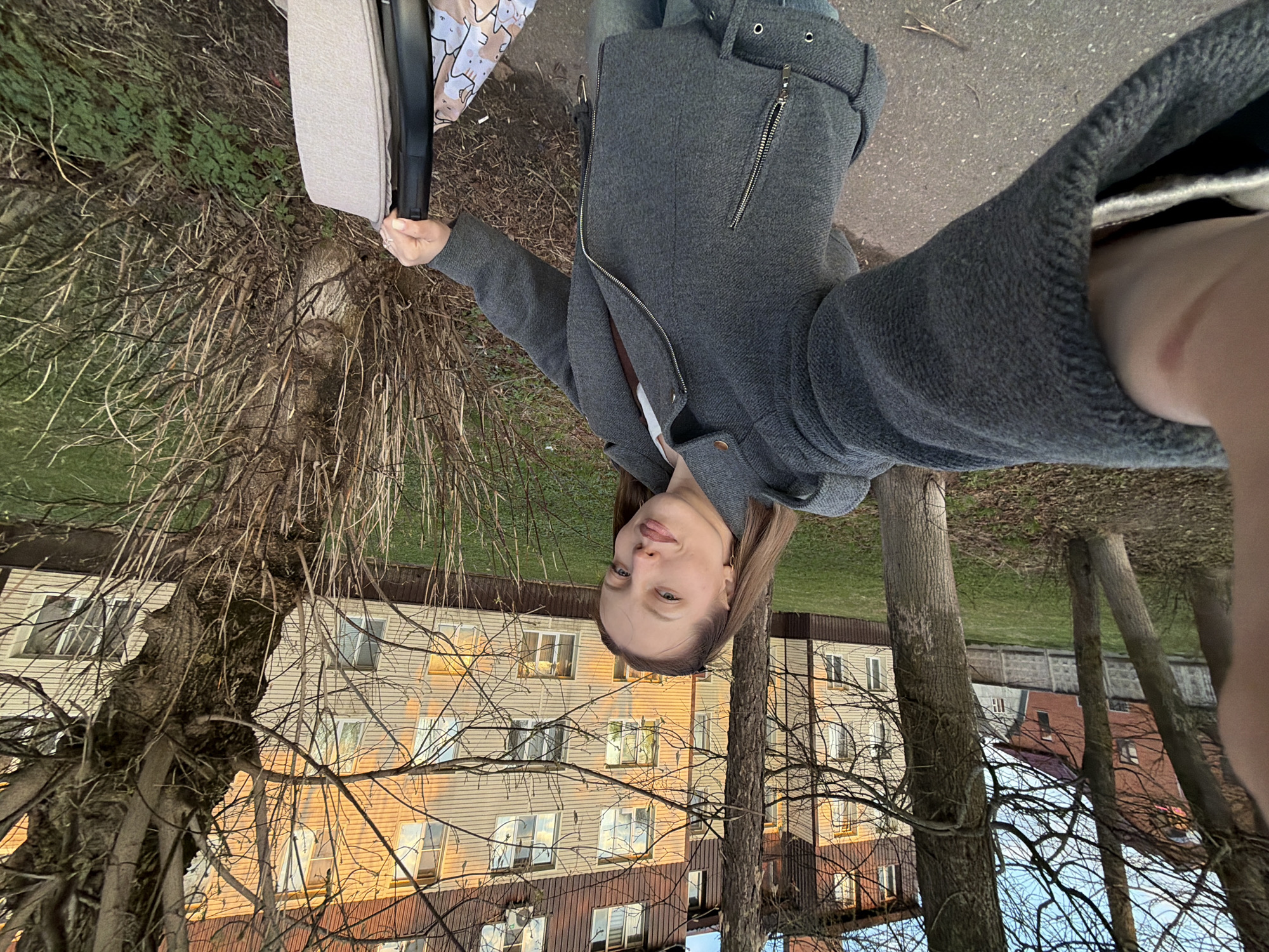 A selfie captures a woman in Reshetnikovo, Russia. She is wearing a gray jacket and holding a baby stroller with a beige interior. The backdrop includes a multi-story building with light-colored siding and a red-brown roof. Trees with bare branches are in the foreground. The sky is visible in the spaces between the trees and building. The image is rotated 180 degrees.