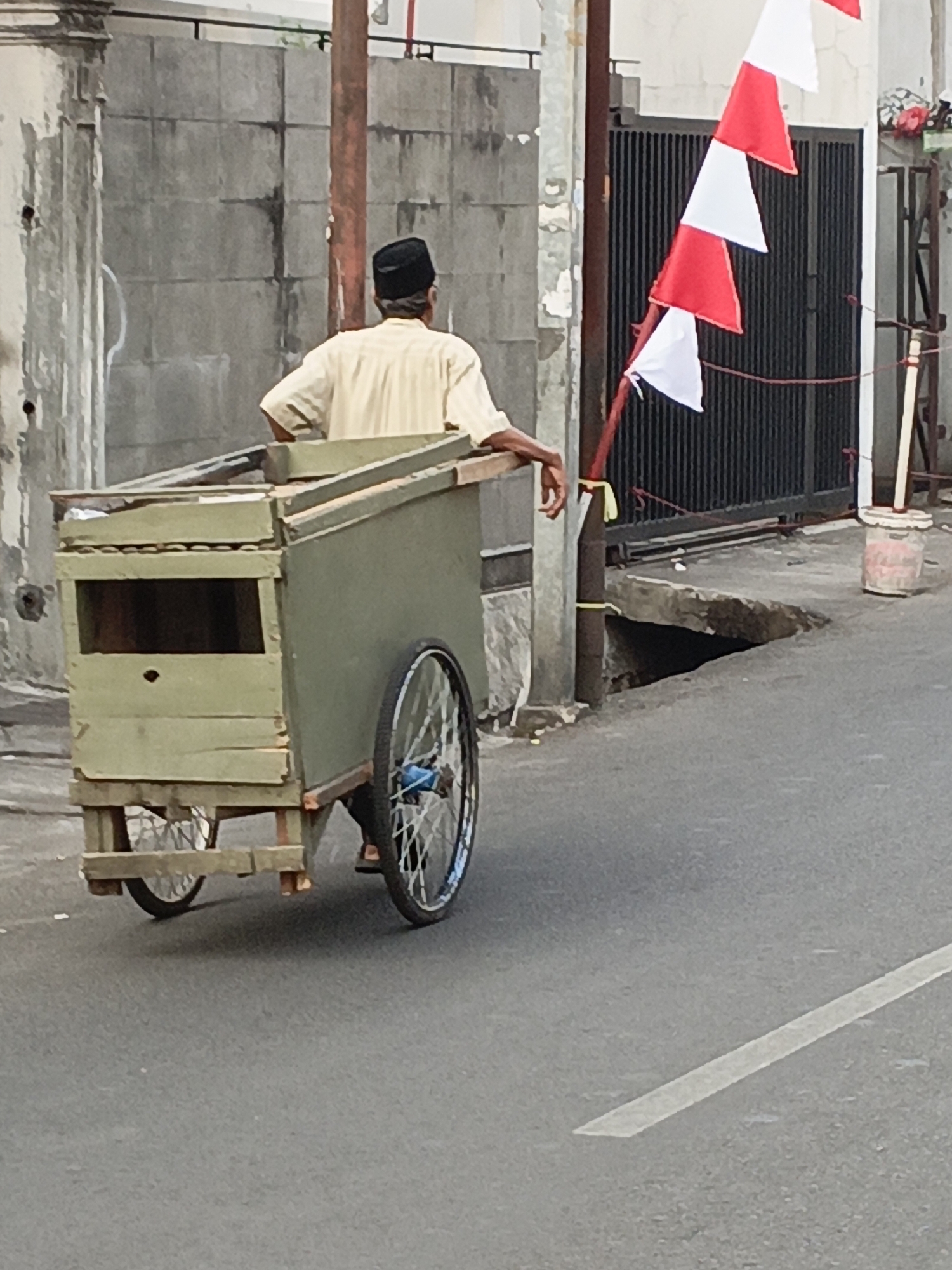 In East Jakarta, Indonesia, a person wearing a black cap and a light-colored shirt is pushing a wooden cart with two wheels on a paved road. The cart is green and appears to be used for transport. A string of red and white triangular flags is displayed on a pole to the right of the cart. The background includes a concrete wall and a black metal gate. There is a visible hole or depression in the ground to the right of the cart, indicating potential uneven terrain. The scene suggests an outdoor setting in a developed area, possibly a street or alleyway.