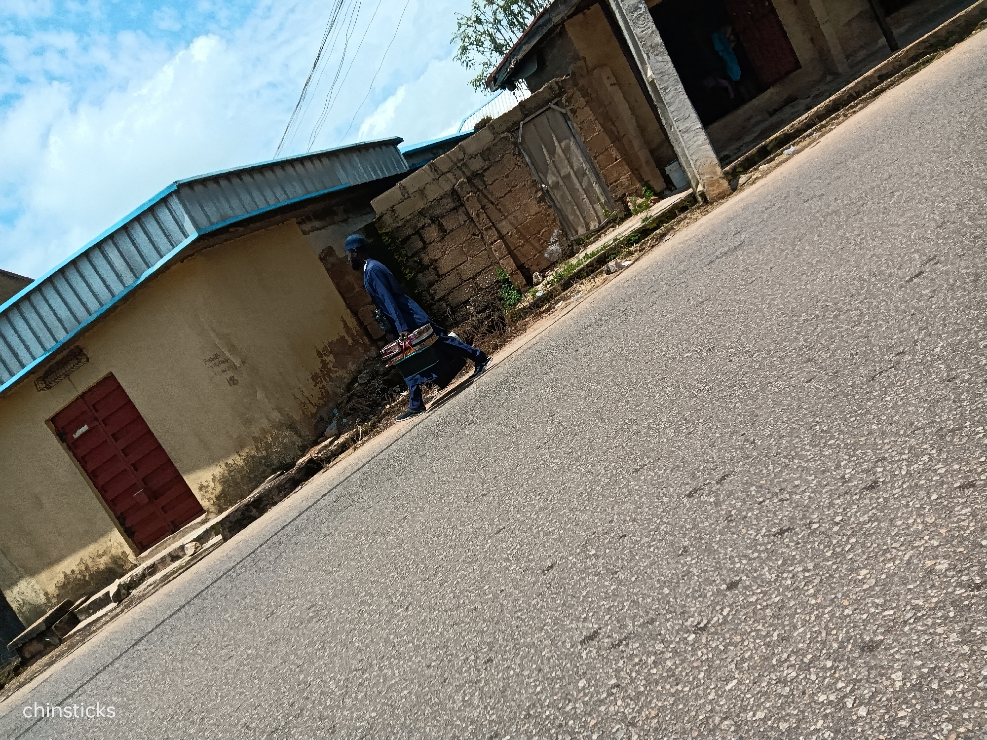 The image depicts a street scene in Jos, Nigeria, with a person walking on the right side of the road, carrying a long, flat object. The person is wearing a blue outfit and a head covering. The street is paved with asphalt, and the right side of the road features a sandy embankment with some sparse vegetation. On the left side of the road, there are buildings with corrugated metal roofs and some brickwork visible. One building has a red metal door. The sky above is blue with scattered white clouds. The photograph is taken at an angle, giving a sense of depth and perspective.