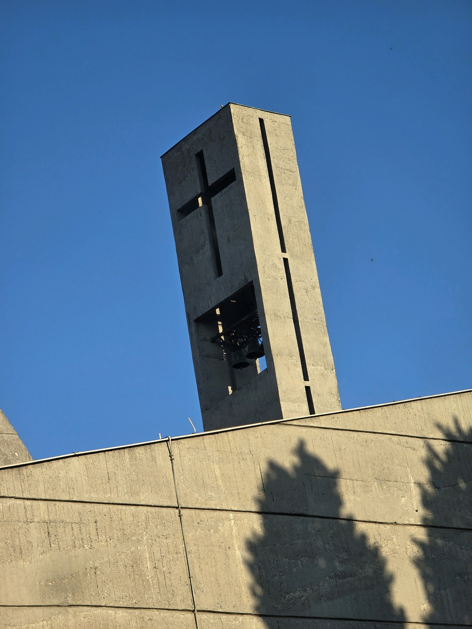 A concrete bell tower, featuring a large cross cut-out and an opening revealing bells, is angled against a clear blue sky. The tower is part of a modern church building, with the top of a concrete wall visible in the foreground. The wall is textured and shows horizontal lines, with the shadows of trees cast across its surface. The overall scene is stark and architectural, with a religious element represented by the cross. The image was taken in Podgorica, Montenegro.