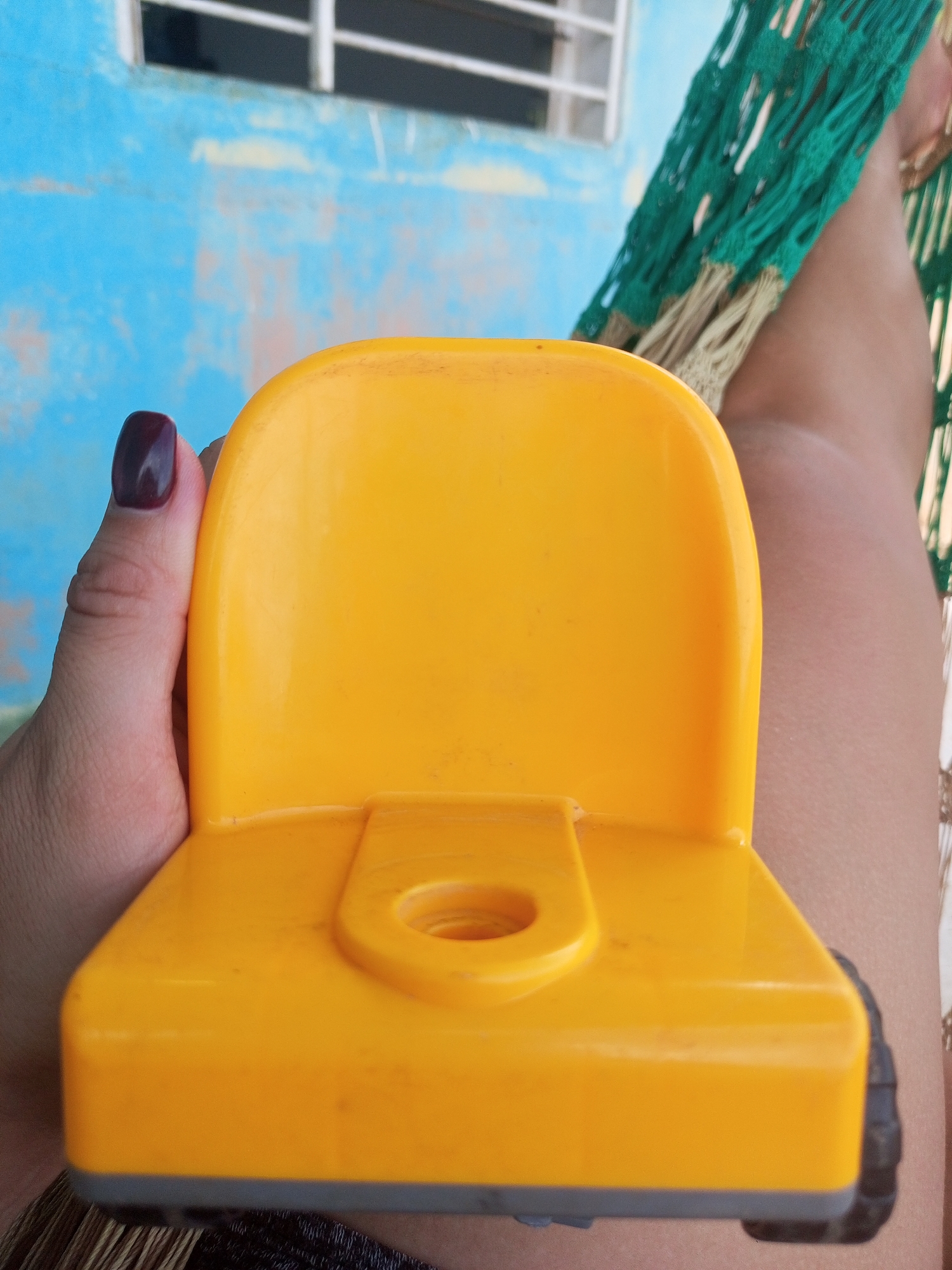 A close-up, eye-level shot captures a person's left hand holding a bright yellow toy truck in La Tagua, Venezuela. The hand, with dark nail polish, is positioned on the left side of the frame, supporting the truck. The toy truck, made of plastic, is yellow with a grey base and black wheels visible on the right side of the image. In the background, a blue wall with a white-framed window and a green hammock are visible. The overall lighting suggests an outdoor setting, possibly during daytime.