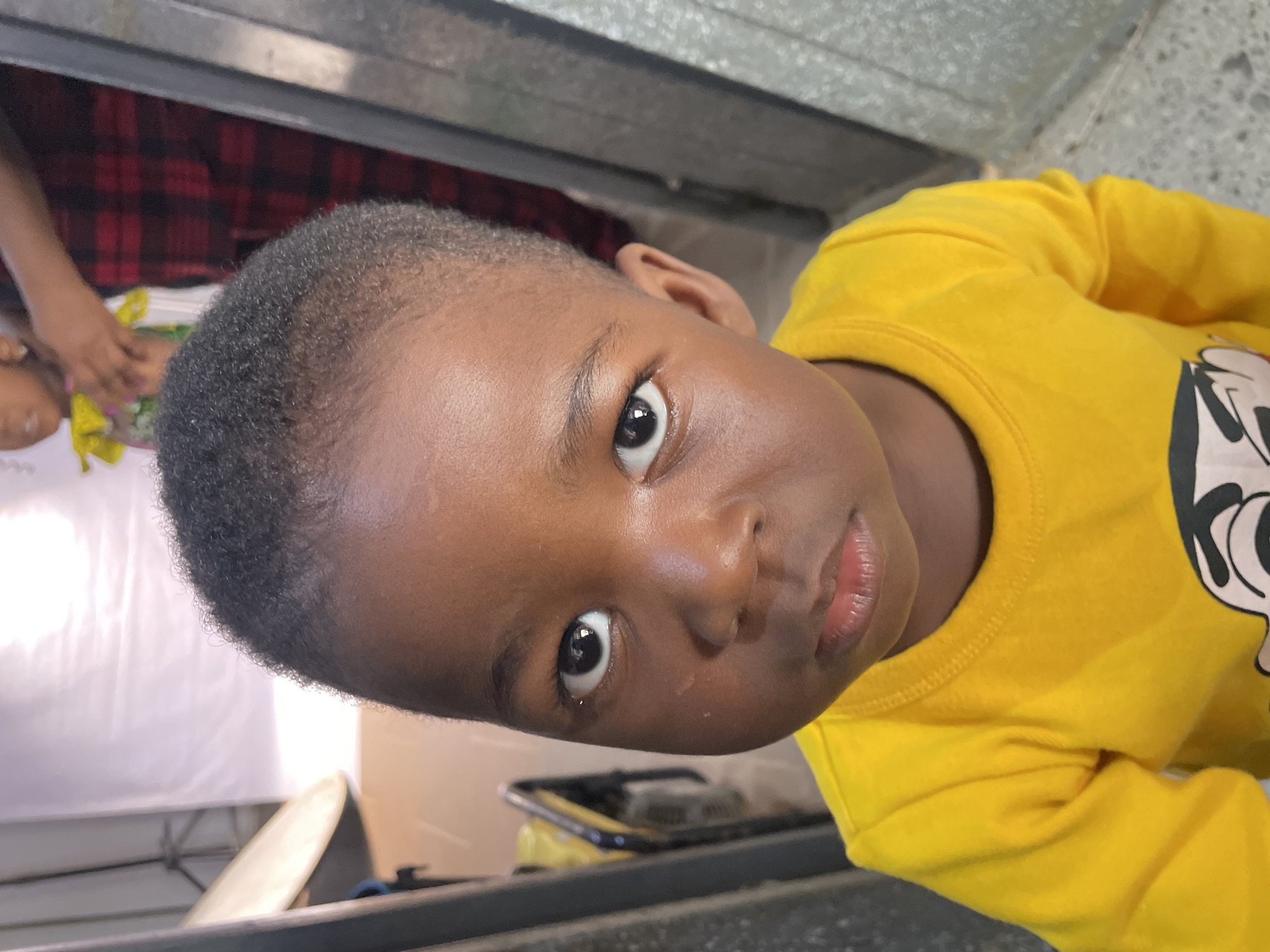 A close-up, tilted shot captures a young, dark-skinned child looking directly at the camera with wide, dark eyes and a calm, engaging expression. The child has short, dark, curly hair and is wearing a bright yellow long-sleeved shirt. A black and white graphic print, possibly a cartoon character's face (resembling Mickey Mouse), is visible on the right side of the shirt.
The setting appears to be indoors. In the blurry background, to the upper left, another person's hand is partially visible, seemingly holding or touching a yellow and green object, possibly a flower or small bouquet. A pattern of red and black plaid fabric can be seen behind the child's head. Various metallic or concrete-like surfaces are present throughout the background, along with a white sheet or backdrop on the far left. On the lower right, a dark rectangular object, possibly a phone or tray, rests on a surface. The overall lighting suggests it is daytime. No discernible text is visible. The scene could be an indoor gathering or event in Jos, Nigeria.