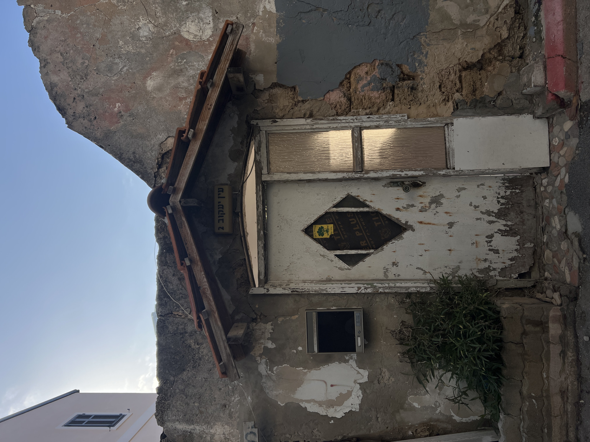 An exterior shot shows a weathered, aged door in Tel Aviv, Israel. The door, painted white and showing significant peeling and wear, features a decorative diamond-shaped insert and a panel of textured glass at the top. A small, framed television is mounted on the wall next to the door, with a patch of green vegetation growing nearby. An overhanging roof structure with clay tiles provides partial shelter. The surrounding wall is heavily textured and in disrepair, with exposed brick and crumbling plaster. A sign in Hebrew is positioned near the roof overhang, and the street borders the right edge.