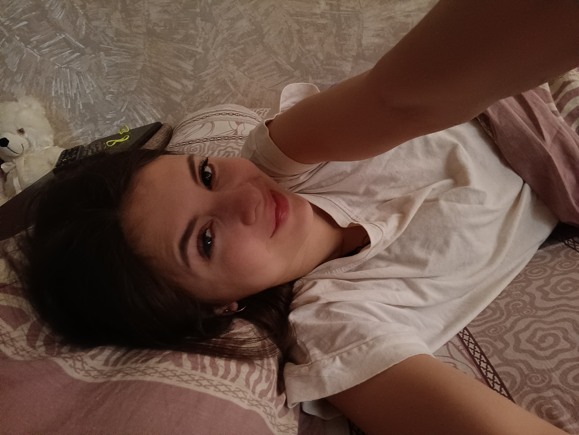 A young woman with dark hair is lying on a patterned bed in a room with textured wallpaper. A stuffed white bear is visible on a surface behind her head, along with a remote control and yellow hair tie. Her face is angled towards the camera with a slight smile. An arm is visible in the upper right corner of the image. The location is Chaikovsky, Russia.