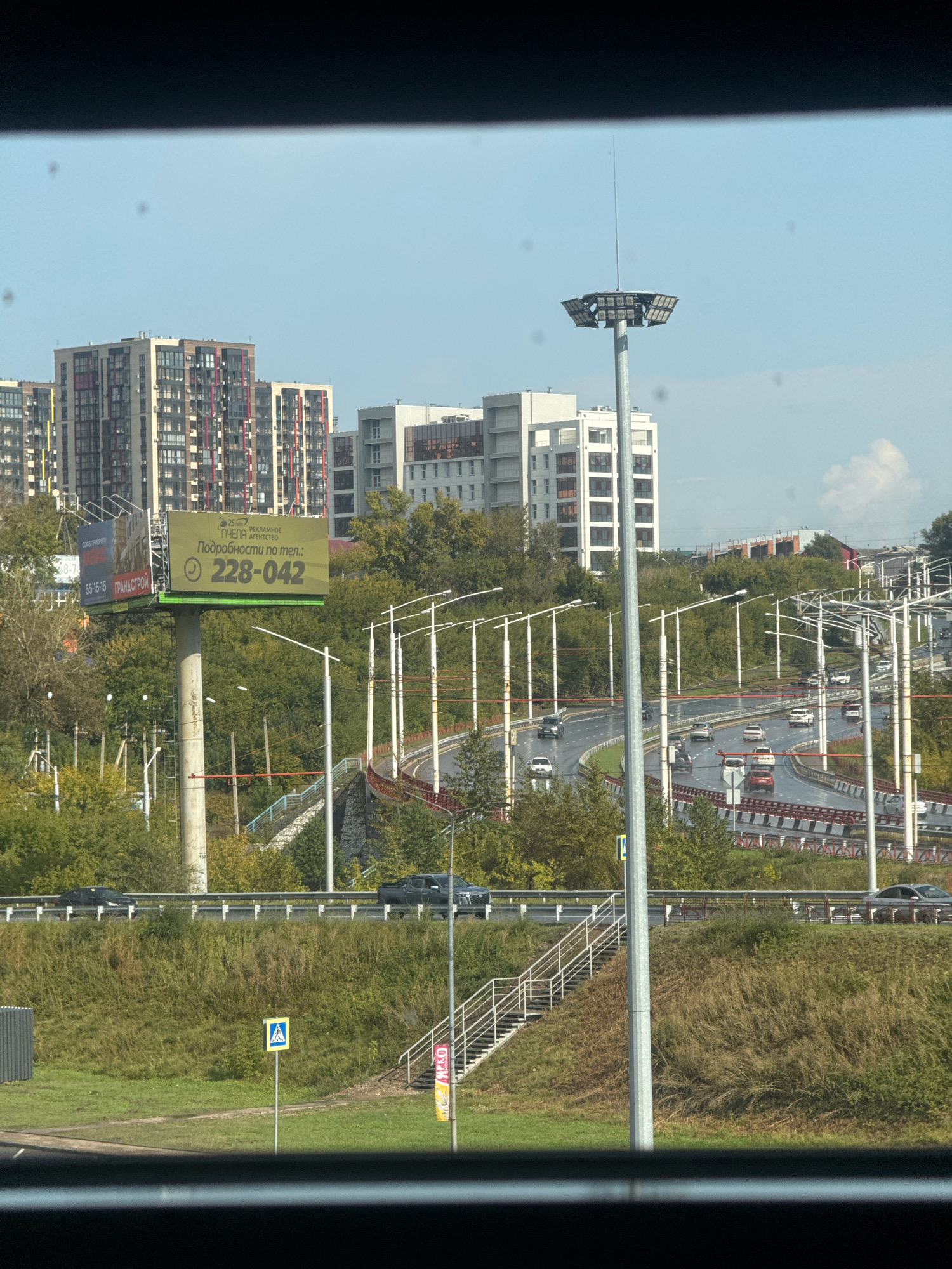 The image depicts a wide, multi-lane road with several vehicles in motion, likely an expressway or highway, surrounded by green, grassy embankments and trees. In the background, there are modern apartment buildings with multiple stories, some featuring colorful accents. A prominent billboard with Russian text and a phone number is visible on the left side. There are numerous streetlights and a tall pole with a lighting fixture in the center of the frame. A pedestrian crossing sign is placed on the grass to the left of a set of stairs leading up a slope. The scene appears to be set in an urban or suburban area of Irkutsk, Russia, on a clear day with a bright sky.
