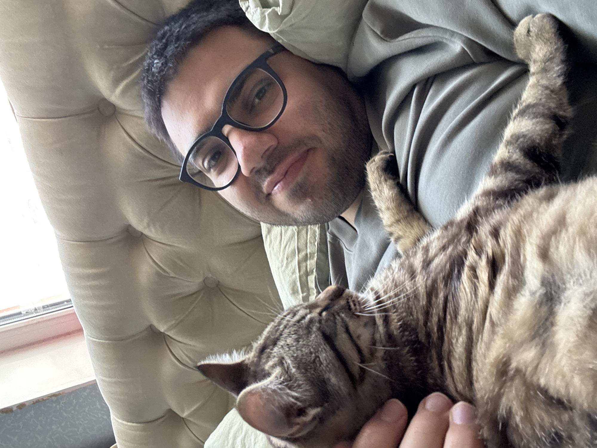 A close-up selfie features a young man with dark hair and a beard, wearing black-rimmed glasses. He is lying down, with his head resting on a tufted, light-colored sofa or bed. Beside him, a tabby cat is snuggled close, with its head resting near the man's face. The man appears relaxed and is looking towards the camera with a soft expression. A hand is visible in the foreground, gently touching the cat. The man is wearing a grey hoodie. The background consists of the sofa and a window with natural light streaming in, suggesting it is daytime. The overall scene evokes a sense of comfort and companionship between the man and his cat. The location is Bila Tserkva, Ukraine. There is no visible text in the image.
