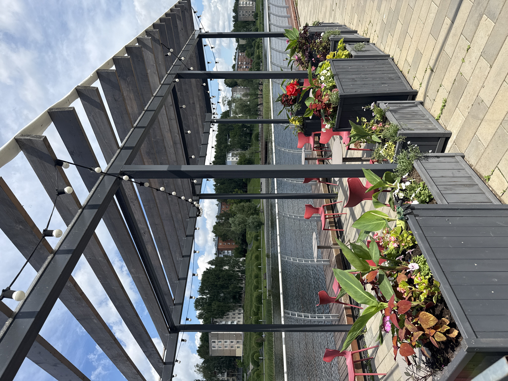 This image depicts an outdoor seating area under a wooden pergola with string lights, situated on a paved walkway next to a body of water. The seating area includes multiple red chairs and small tables. In the foreground, several dark gray planters are filled with a variety of plants, including large green leaves, red and green foliage, and small pink and white flowers.
In the background, across the water, is a green, hilly landscape with several multi-story residential buildings and trees. The sky is partly cloudy and blue. The walkway is paved with rectangular stones laid in a diagonal pattern.
The scene appears to be in the city of Votkinsk, Russia, a location known for its picturesque waterfront areas and recreational spaces. The overall impression is that of a pleasant outdoor cafe or restaurant setting by the water. There are no people clearly visible in the image, but the setup suggests a place for public gathering and dining.