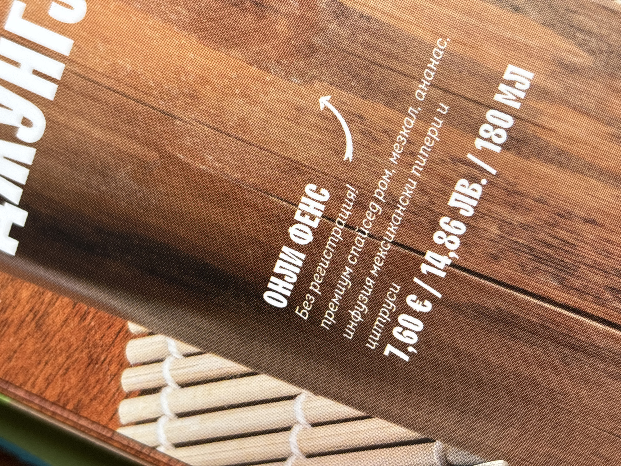 The image shows part of a promotional menu for "OHLI FENCE", a drink made without registration. The drink consists of premium spiced rum, mescal, pineapple, and an infusion of Mexican peppers and citrus. The price is 7.60€ / 14.86 лв. / 180 ml. The image depicts a close-up of the printed menu. The image background suggests the location is somewhere that serves this drink, possibly a restaurant, bar or club.