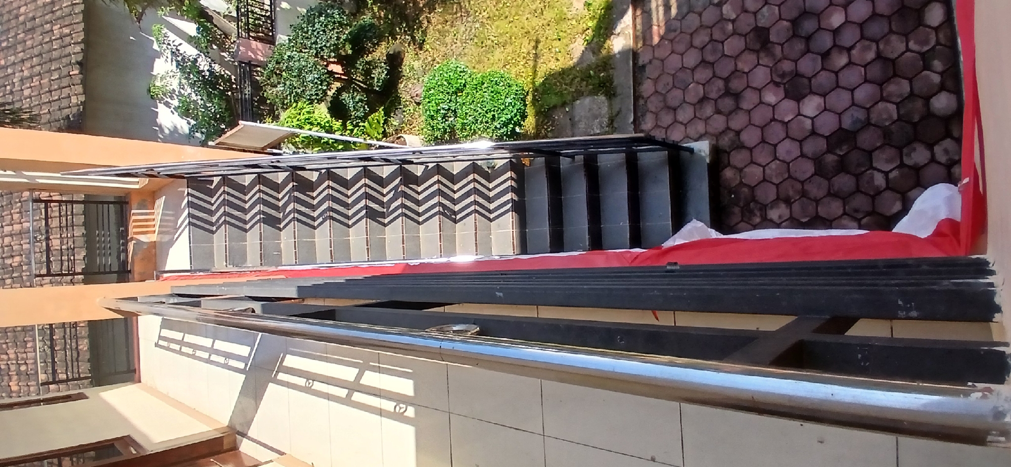 The image shows an outdoor staircase with a zigzag shadow pattern cast across its steps. The staircase is made of dark tiles and has a black metal railing on the right side. A red and white flag is draped along the side of the staircase. To the left of the staircase is a tiled walkway with long shadows. In the background, there are trees, grass, and hexagonal paving stones. The scene appears to be a building exterior, possibly in a residential or institutional setting. The location is described as the city of Pejaten, Indonesia, which is a subdistrict in Pasar Minggu, South Jakarta, Indonesia.