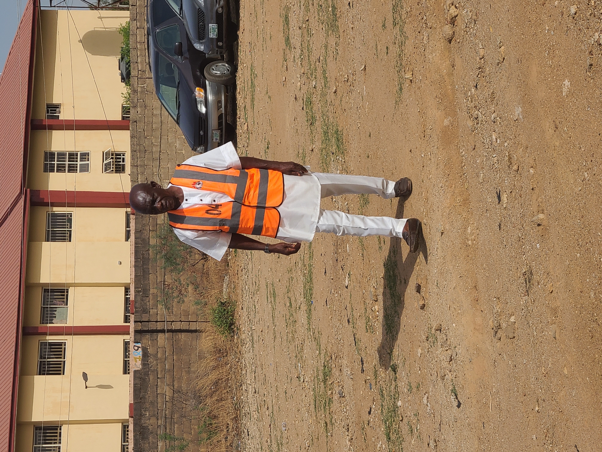 The image shows a man in Jos, Nigeria, wearing an orange reflective vest over a white outfit and brown shoes. He is standing on a dirt and gravel ground. Behind him is a light-colored building with red trim and windows, and a dark vehicle. The location appears to be an outdoor area with limited vegetation.