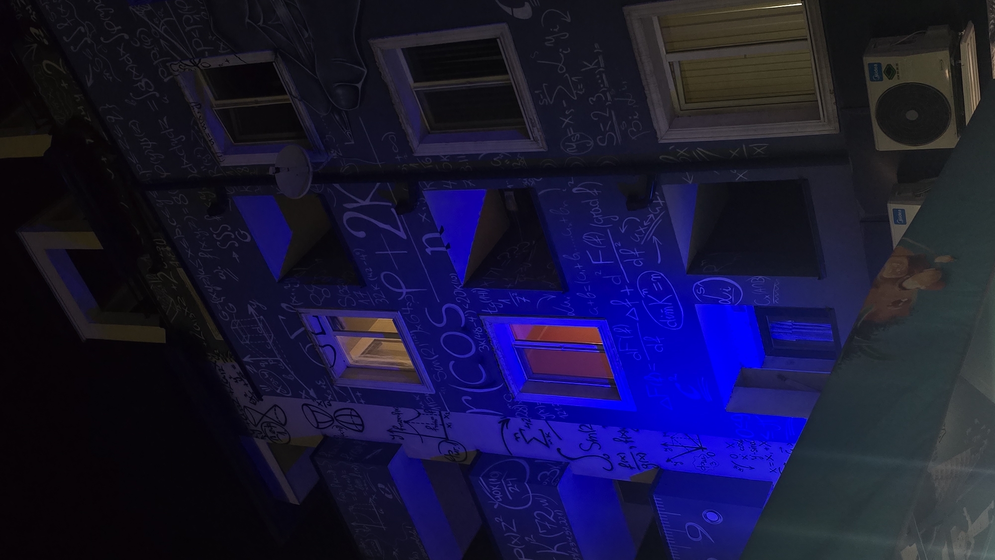 The image shows a building facade in Sofia, Bulgaria, adorned with mathematical equations and formulas. The building is painted in a deep blue color, and the equations are written in white. Several windows are visible, some illuminated from within, and air conditioning units are mounted on the building's exterior. The lighting is a mix of artificial and natural, with blue light highlighting certain parts of the structure. A satellite dish is also attached to the building. The overall impression is that of a building celebrating mathematics or science.