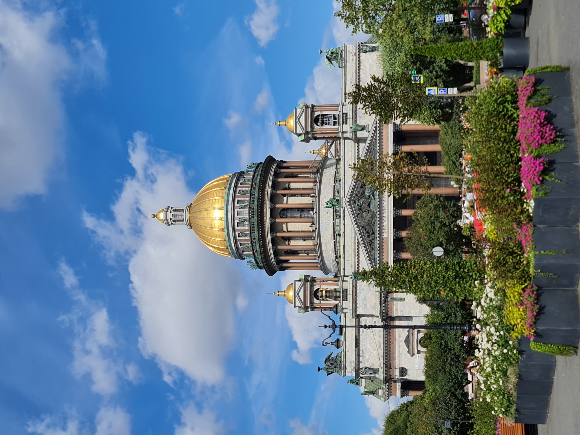 The image depicts Saint Isaac's Cathedral in Saint Petersburg, Russia. The grand cathedral, with its iconic golden dome, stands tall against a vibrant blue sky dotted with white clouds. In the foreground, a well-maintained flower bed is filled with a colorful array of blooming plants, including white and pink flowers. Several dark gray planters line the flower bed, adding a modern touch to the scene. Behind the flowers, trees and shrubbery provide a lush green backdrop. A lamppost is visible to the left of the cathedral. People are faintly visible in the background, suggesting a public space or park setting. The overall impression is one of architectural grandeur and natural beauty.