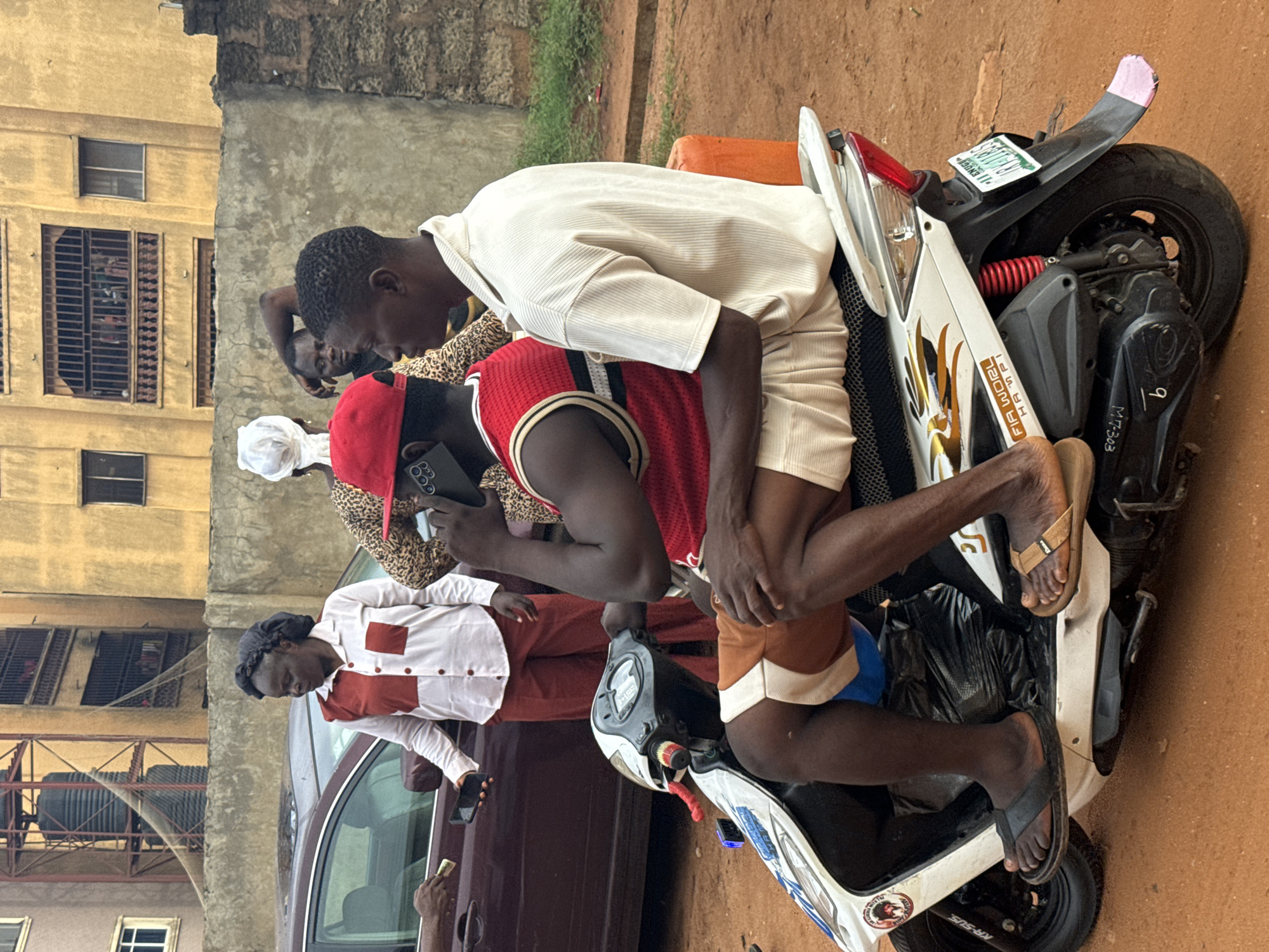 An outdoor scene in Onitsha, Nigeria, captures a dynamic street interaction during daytime. The image, rotated 90 degrees counter-clockwise, shows a group of people gathered around a parked motorcycle and car on an earthy, reddish-brown ground, against a backdrop of multi-story buildings.
In the foreground, a white and black scooter is prominent. A dark-skinned man in brown shorts and a red sleeveless jersey with black and white trim sits on the scooter, looking down at his phone. Another dark-skinned man, wearing a light beige short-sleeved shirt and light-colored shorts, leans over the first man, looking down as well. Both are wearing flip-flops. The scooter features gold-colored decals, a red suspension spring, and a black seat. Visible text on the scooter includes "FIA WORL HA API" (partially obscured, possibly "FIA WORLD HAPPY") and "KR-SUS" near the front wheel. The rear fender displays a green Nigerian license plate with "LAGOS, THE CENTRE OF EXCELLENCE" and the alphanumeric code "LSF-676XA" (partially visible). Another marking "MT-303" is on the side.
To the left, a dark-colored car is parked. A dark-skinned woman with hair covered by a dark net stands beside the car, wearing a white long-sleeved shirt with rust-colored rectangular patches on the chest and matching rust-colored trousers. She holds a phone in her hand. A hand from inside or near the car extends, holding folded currency notes towards her, suggesting a transaction.
In the background, other individuals are partially visible: one person wearing a white headscarf and a leopard-print top, and another dark-skinned person further back. The backdrop consists of older, multi-story buildings, one yellowish and another grey, with barred windows. The overall atmosphere appears casual, possibly depicting a common street scene or informal transaction in an urban environment.