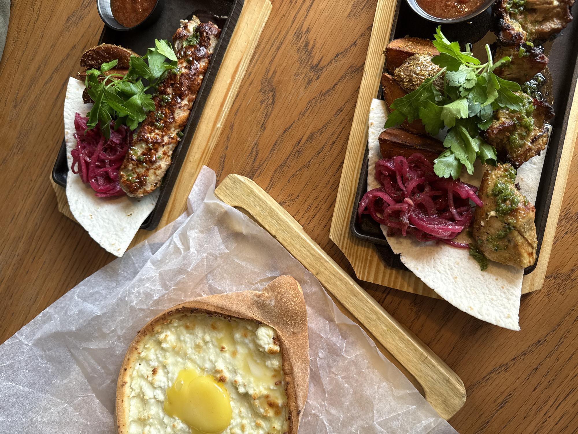 This overhead image captures a spread of traditional Caucasian or Georgian dishes laid out on a polished wooden table, likely in a restaurant in Votkinsk, Russia. The scene depicts a hearty meal ready to be enjoyed.
Two black rectangular platters, possibly cast iron, are presented on light wooden serving boards with raised edges. The platter on the left features a long piece of grilled minced meat, resembling a lulya-kebab, generously garnished with fresh green herbs, and served alongside vibrant pickled red onions and golden-brown potato wedges. A small black bowl containing a reddish sauce is positioned in the upper left corner of this platter, all resting on a piece of soft flatbread.
The platter on the right holds several pieces of grilled meat, akin to shashlik (skewered meat), coated with a green sauce or herb mixture. It is similarly accompanied by pickled red onions, fresh green herbs, and potato wedges, with a small bowl of sauce and a flatbread underneath.
In the foreground, placed on white parchment paper, is a freshly baked Adjaruli Khachapuri. This boat-shaped bread is filled with melted white cheese, with a bright yellow, runny egg yolk centered in the middle. A light wooden utensil or part of a serving tray is visible between the khachapuri and the right platter.
The lighting is bright and even, indicative of an indoor dining setting. The food's presentation is rustic yet appealing, highlighting the textures and colors of the ingredients. No people or text are visible in the image.