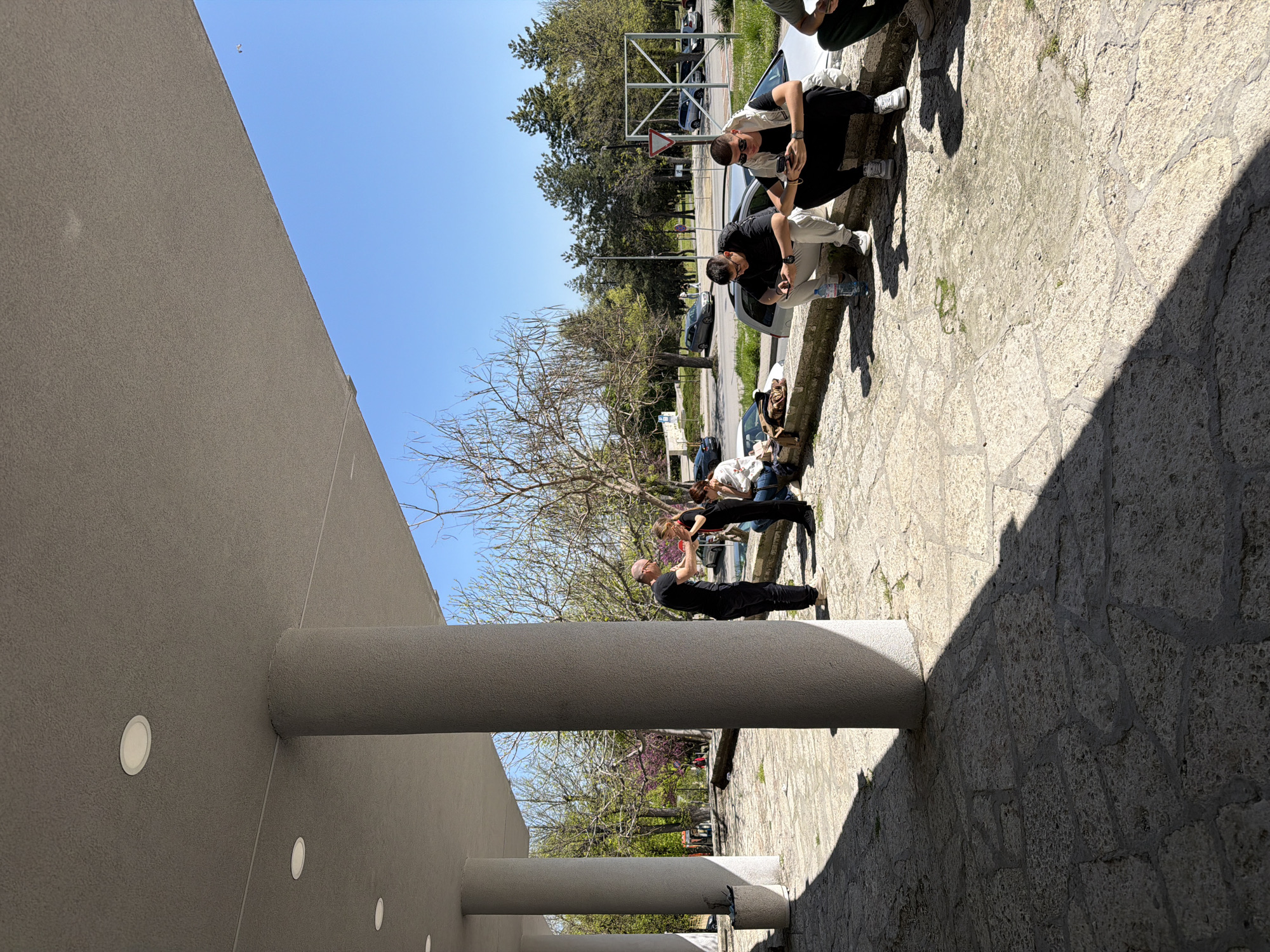 The image is taken at an outdoor location in Shumen, Bulgaria. It shows a group of people interacting near a building with cylindrical column-like architectural features. The ground is paved with light-colored stone. Several trees are visible in the background. Some of the people appear to be inspecting a vehicle parked nearby. One individual seems to be taking pictures of another. The day is sunny, with shadows cast by the building and columns.