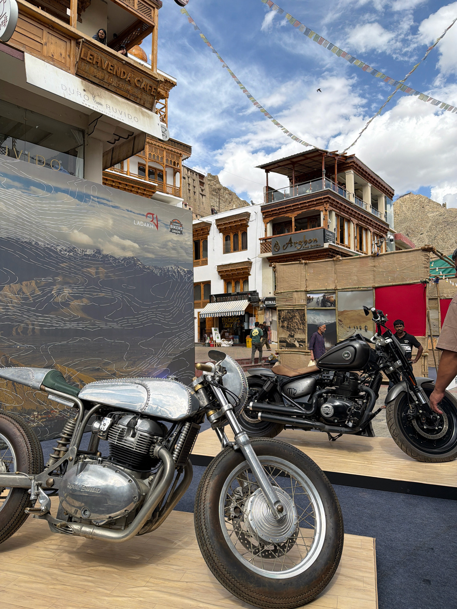 In Leh, India, two Royal Enfield motorcycles are displayed on a wooden platform in front of a building with traditional Ladakhi architecture. The building features multiple levels with wooden balconies and decorative latticework. A banner displaying a topographical map of Ladakh and the "Ladakh Bikers Union" logo is visible behind the motorcycle in the foreground. The motorcycles appear to be custom-built. The foreground motorcycle is a chrome-finished cafe racer, while the motorcycle behind it is a matte black cruiser. In the background, people are seen walking on the street and interacting with vendors. Prayer flags are strung across the sky, adding a cultural element to the scene. The sky is partly cloudy with visible mountains in the distance, indicating the characteristic landscape of Ladakh.