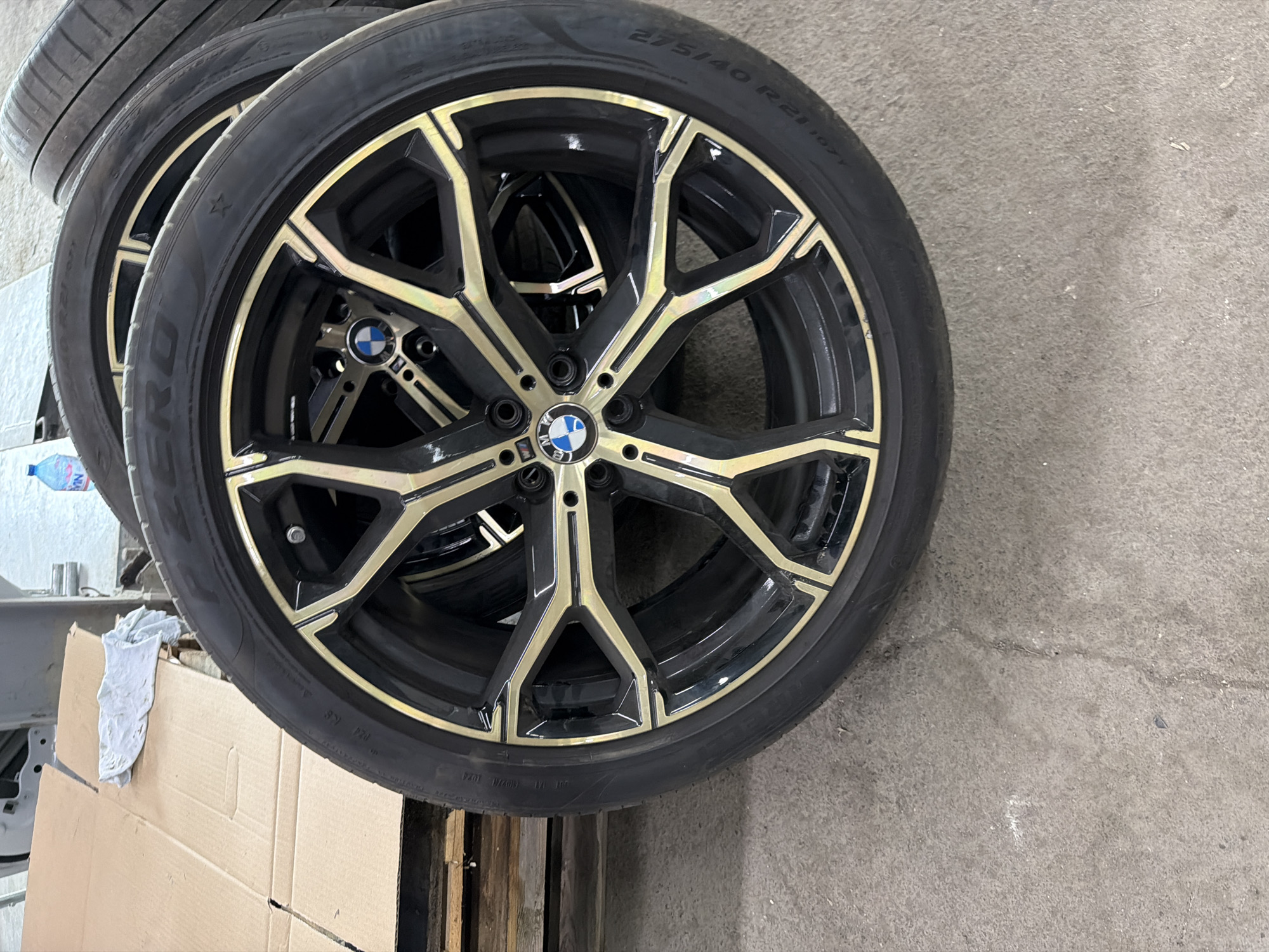 The image displays a collection of automotive wheels and tires, likely in a garage or workshop setting. The primary subject is a large, black multi-spoke wheel with prominent gold or bronze-colored accents. A BMW logo is clearly visible on the central hubcap, and a smaller "M" logo is present on one of the lug nut covers, indicating an M Performance rim. The tire mounted on this rim bears the size specification "275/40 R21 107Y" and a star symbol, suggesting it's an original equipment tire for BMW.
Several other similar wheels are stacked behind the prominent one, partially visible, suggesting either a set or inventory. The setting features a concrete floor, which takes up a significant portion of the right side of the frame. In the background to the left, there are cardboard boxes, what appears to be a wooden pallet, and a white or light-colored wall. A plastic water bottle with "HANA" visible on its label rests near the wall. The overall activity depicted is the storage or display of these automotive components. The lighting is consistent with an indoor industrial environment, and no people are present.