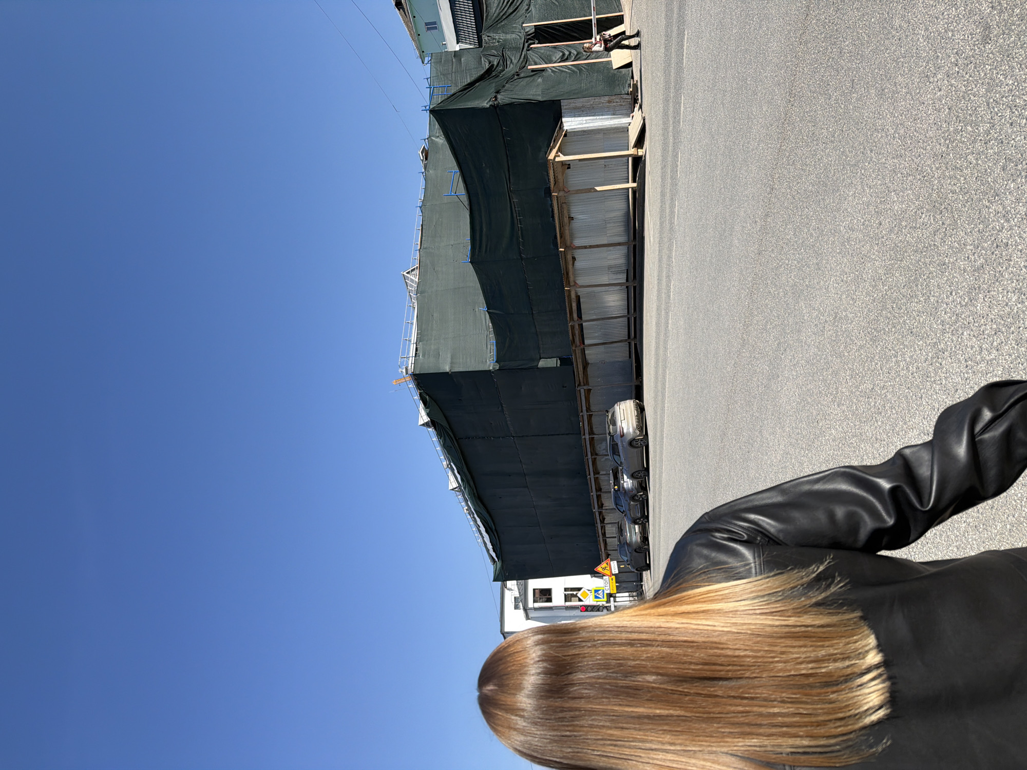 A daytime street scene in Perm, Russia shows a building under construction with scaffolding covered in a dark green screen. A paved road runs alongside the building with parked cars partially visible under the scaffolding. A person wearing a black leather jacket is partially visible in the foreground, with light brown hair extending from the top of the head. Street signs are present near the construction site, and the sky is clear and blue.