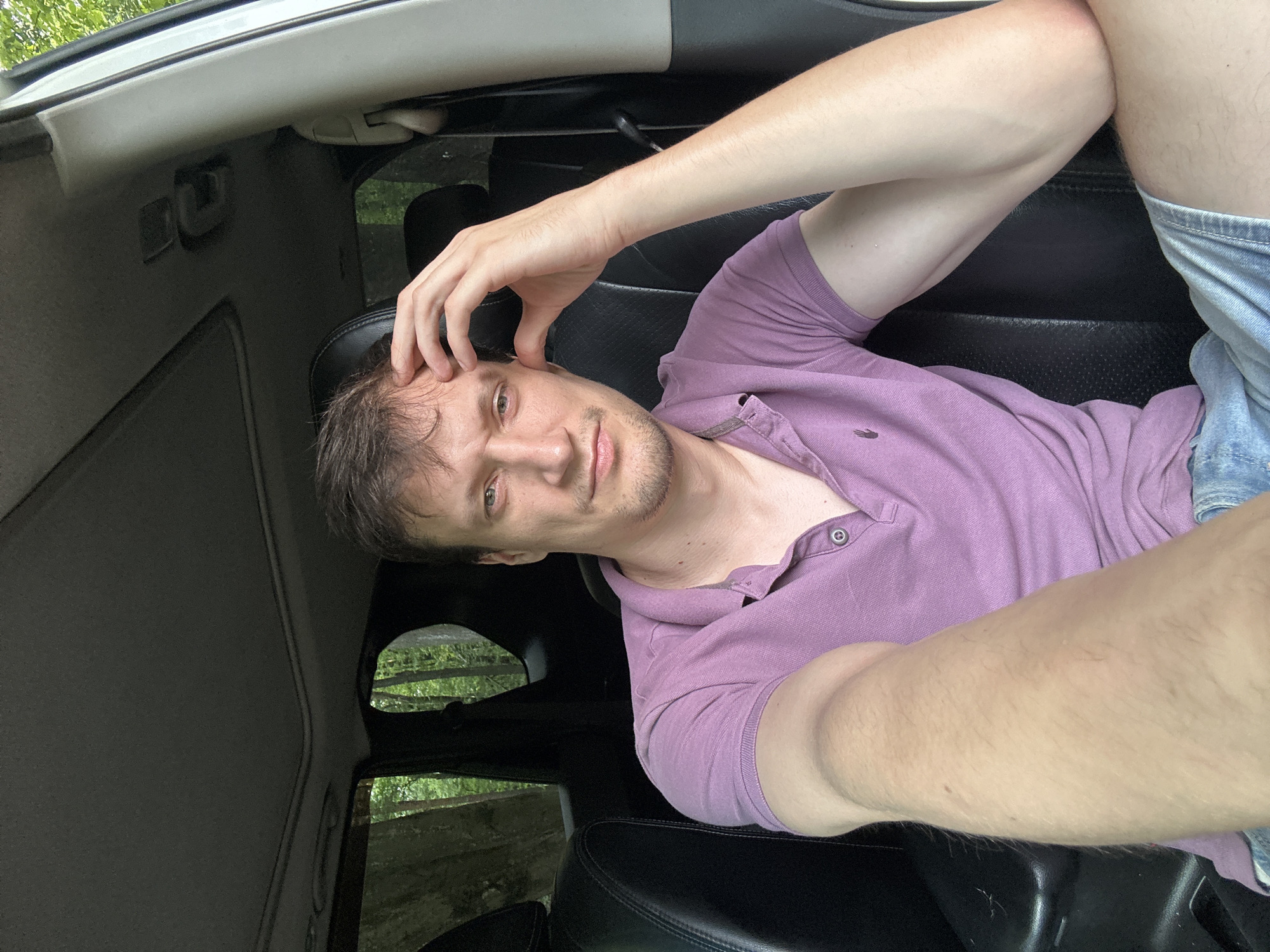 A medium shot captures a man sitting in the driver's seat of a vehicle, with his right hand touching his forehead. The man, who appears to be in his late 20s or early 30s, has short brown hair and is wearing a purple polo shirt and denim shorts. The interior of the car, with its black leather seats and sunroof, is visible. Through the car windows, a lush green forest can be seen, suggesting a rural or natural setting. The image does not contain any information to indicate the location in Shatava, Ukraine.