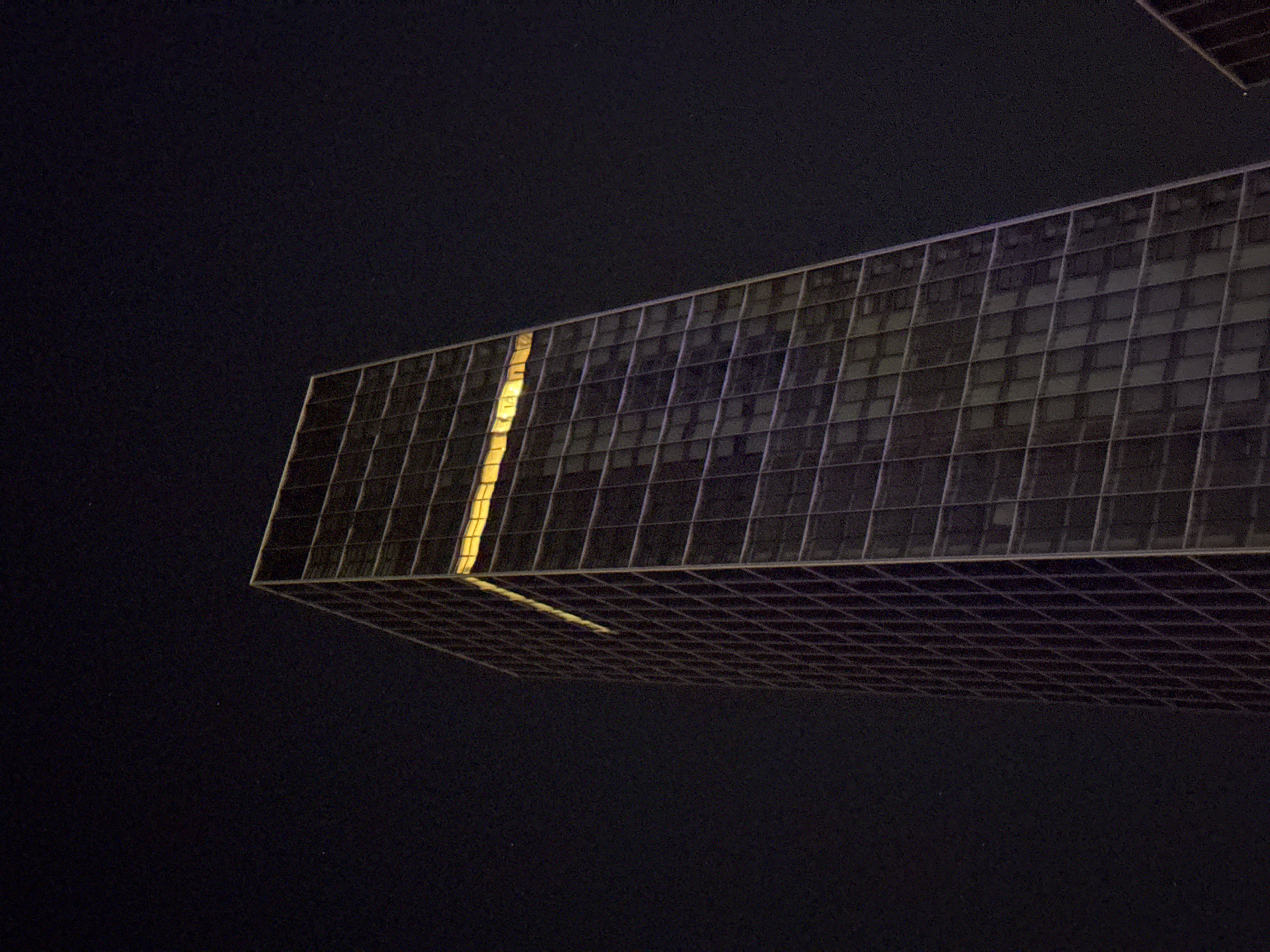 The image displays a modern architectural building against a dark night sky, suggesting a clear night. The primary subject is a large, rectangular section of a glass-facade building, viewed from an unconventional low angle, creating the illusion of it projecting or hovering against the black background.
The building's surface is composed of numerous rectangular glass panels divided by a grid of structural framing. A prominent, vertical strip of bright yellow light emanates from within one section of the building, standing out vividly against the otherwise dark or dimly lit facade. This light source is reflected on the visible underside or a lower reflective surface of the building, forming a fainter, parallel yellow line along its grid structure.
The background is uniformly dark, indicative of a night sky, and exhibits a noticeable fine grain or digital noise. No human figures or discernible activities are present. The image focuses entirely on the striking geometry of the building and the stark contrast between the internal light and the surrounding darkness. There are no specific location cues visible within the photograph, but the provided context places the scene in Perth, Australia. No text is visible in the media file.