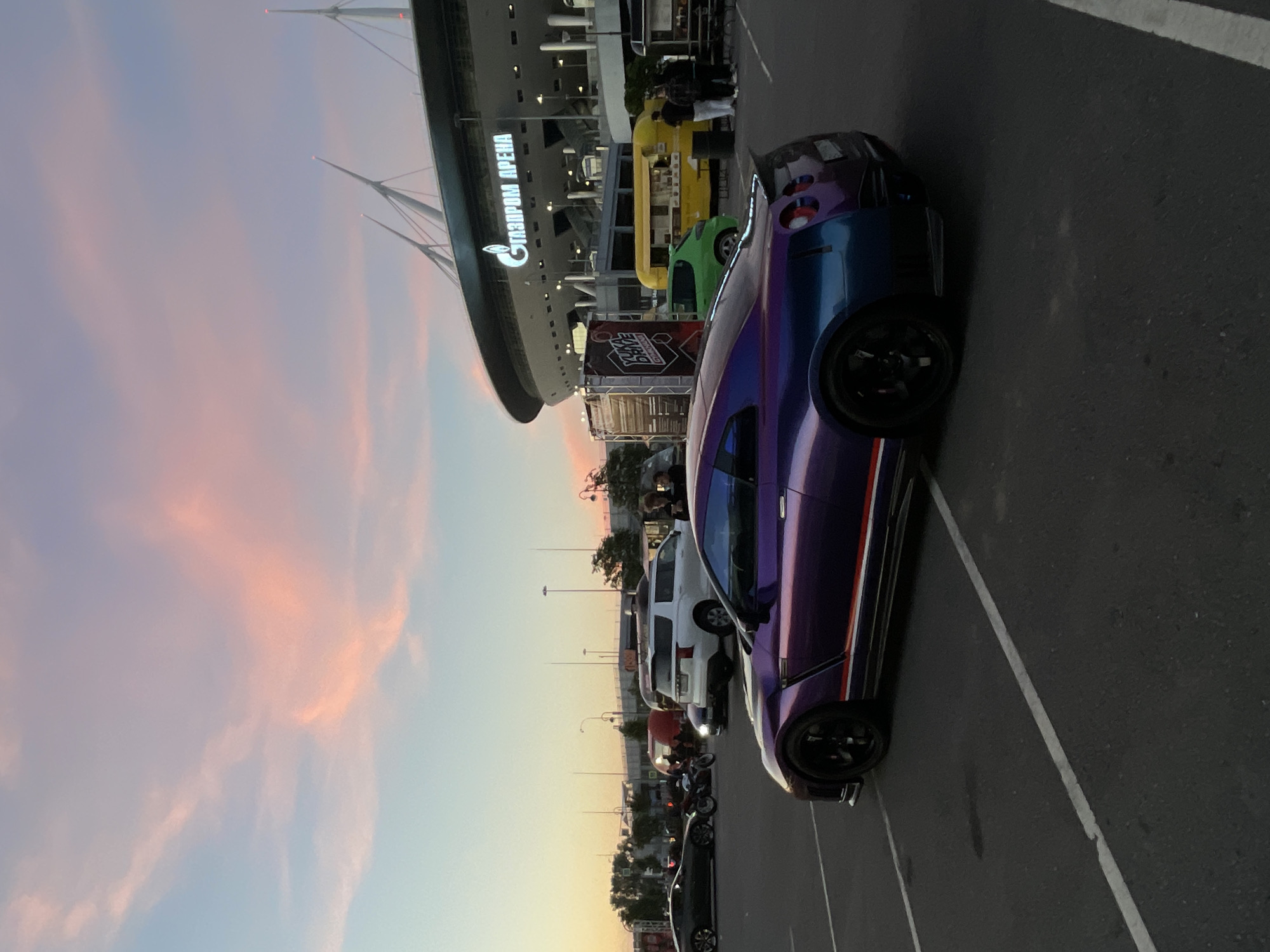 A purple and blue Nissan GT-R is parked in a lot in front of the Gazprom Arena in St. Petersburg, Russia. The sky is painted with hues of pink, orange, and blue as the sun sets. Several other vehicles, including a white SUV and a green compact car, are visible in the background, along with food trucks and a tent. The stadium's distinctive illuminated sign reads "GAZPROM ARENA." A group of people are gathered near the food trucks. The lighting suggests it is dusk.
