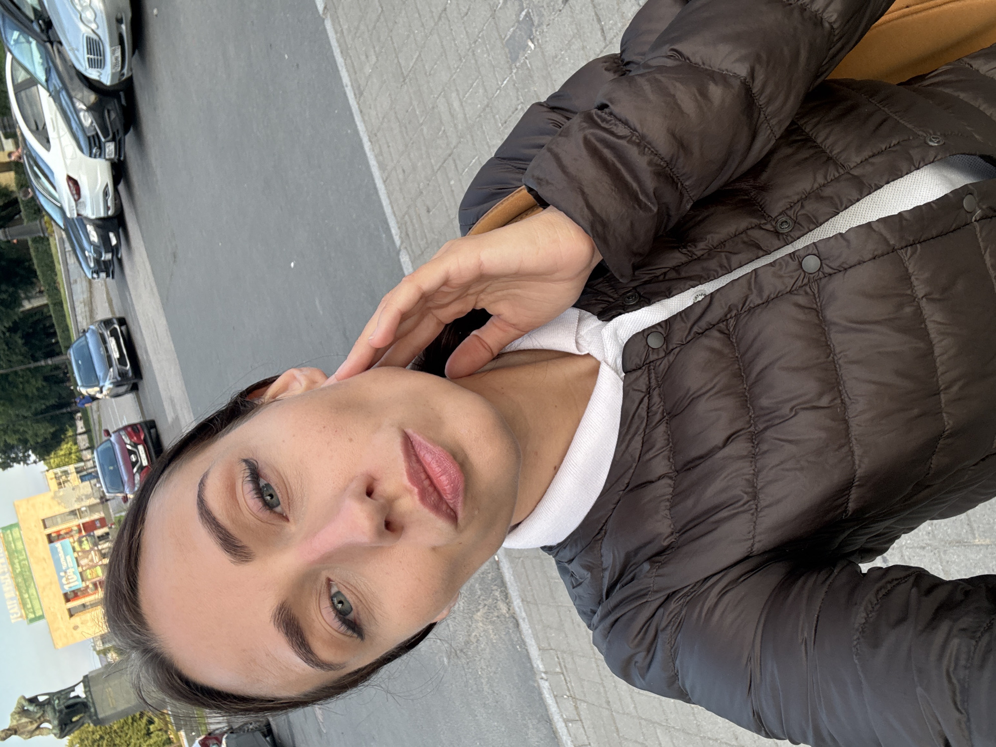 The media file is a selfie taken outdoors in what appears to be a city environment, likely Saint Petersburg, Russia, given the text on a building in the background. The subject is a woman with dark hair and light-colored eyes, wearing a brown puffer jacket over a white collared shirt. She is holding her hand up to her face, with her fingers gently touching her cheek and neck. Her expression is neutral or slightly pensive.
The background reveals a street scene with several cars parked along the road. There are also some buildings and trees visible. One building in the mid-ground has a large sign with text in what appears to be Cyrillic script. A statue or monument is also visible in the lower left portion of the frame. The pavement is made of cobblestones or similar material. The lighting suggests it is daytime, with possibly overcast or diffused sunlight. The overall impression is a candid moment captured during a casual outing.
