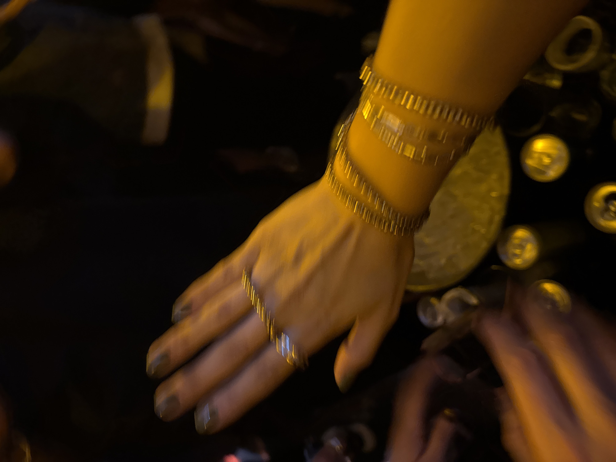 The image shows a hand with several gold bracelets on the wrist and a gold ring on the finger. The nails are painted a dark color. The hand appears to be reaching towards a table surface that is covered in round, metallic objects that could be bottle caps or coins. Another hand is partially visible in the lower right corner. The image is blurred, and the scene is dimly lit. There is no information in the image that indicates its location is in Mexico City, Mexico.