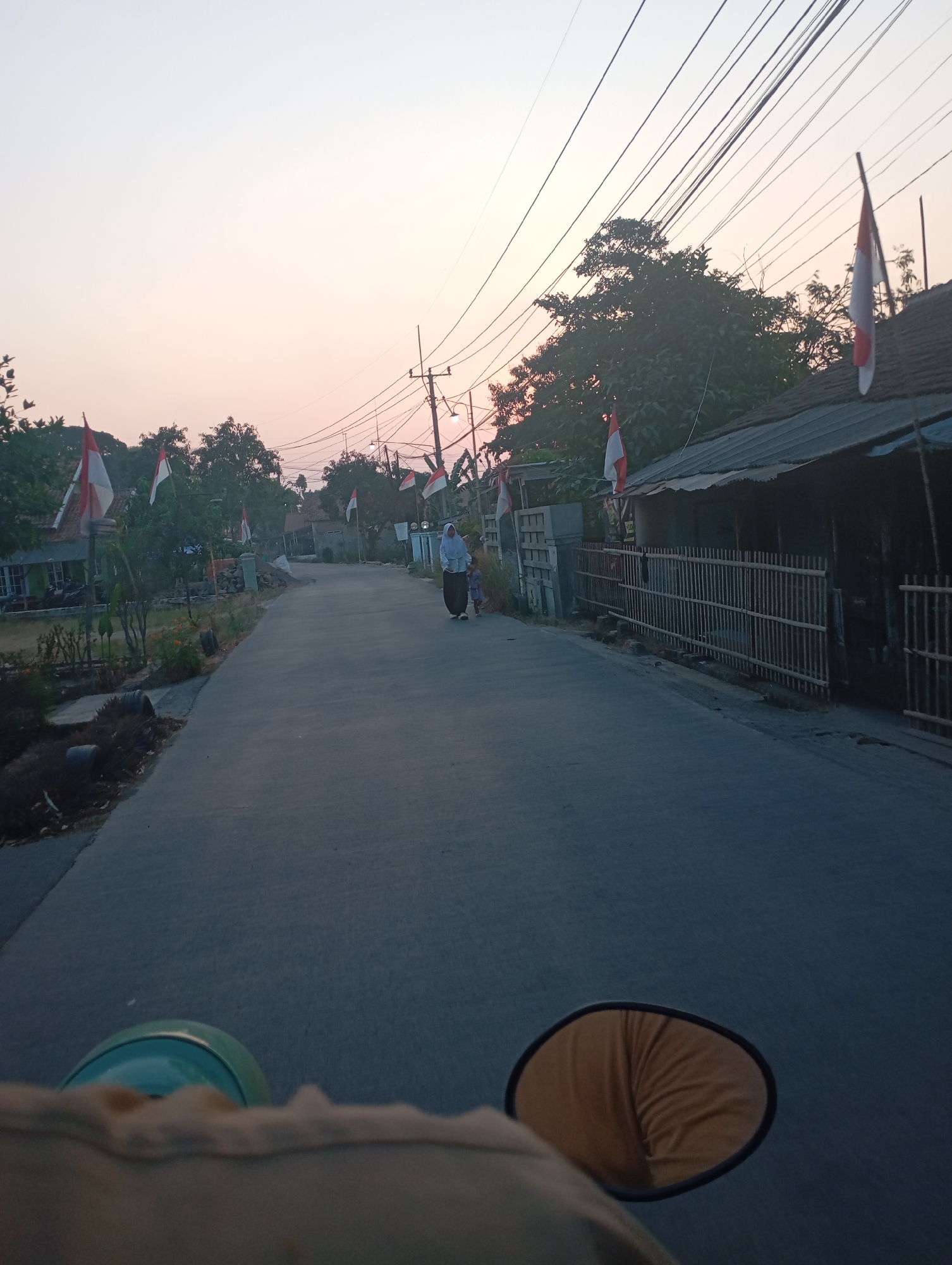 The image depicts a paved road in a residential area during the late afternoon or early evening, as indicated by the soft, diffused light and the pastel colors in the sky. The scene is likely located in Kecamatan Haurgeulis, Indonesia, given the presence of Indonesian flags. The road is lined on both sides by houses and trees. On the left side of the road, there are houses with a small garden area. Several Indonesian flags, red and white, are prominently displayed on poles along the road, suggesting a national celebration or commemoration. On the right side of the road, there is a row of houses with a traditional bamboo fence in front of them. A woman and a child are walking away from the camera on the right side of the road. The foreground of the image is dominated by the perspective from a vehicle, likely a motorcycle, showing a portion of its handle and side mirror. The overall atmosphere is serene and rural.