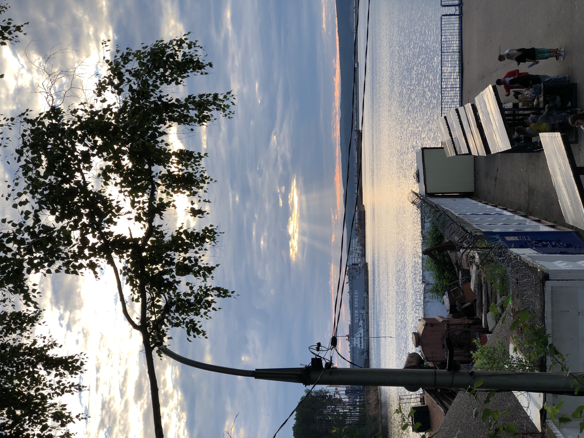 The image shows a view of the Kama River in Chaikovsky, Russia, during a sunset. In the foreground, a silhouetted tree branch with leaves hangs over the scene, with a lamp post on the left. Along the riverbank, there are industrial elements including a barge or ship with "SLOW SPEED!" visible on its side, a fenced-off area with rubble, construction materials, and a small structure. Further down the riverbank, a group of people are gathered under a series of slanted, reflective structures, possibly solar panels, and are interacting with each other. The sky is cloudy with rays of sunlight breaking through, casting a warm glow on the water.
