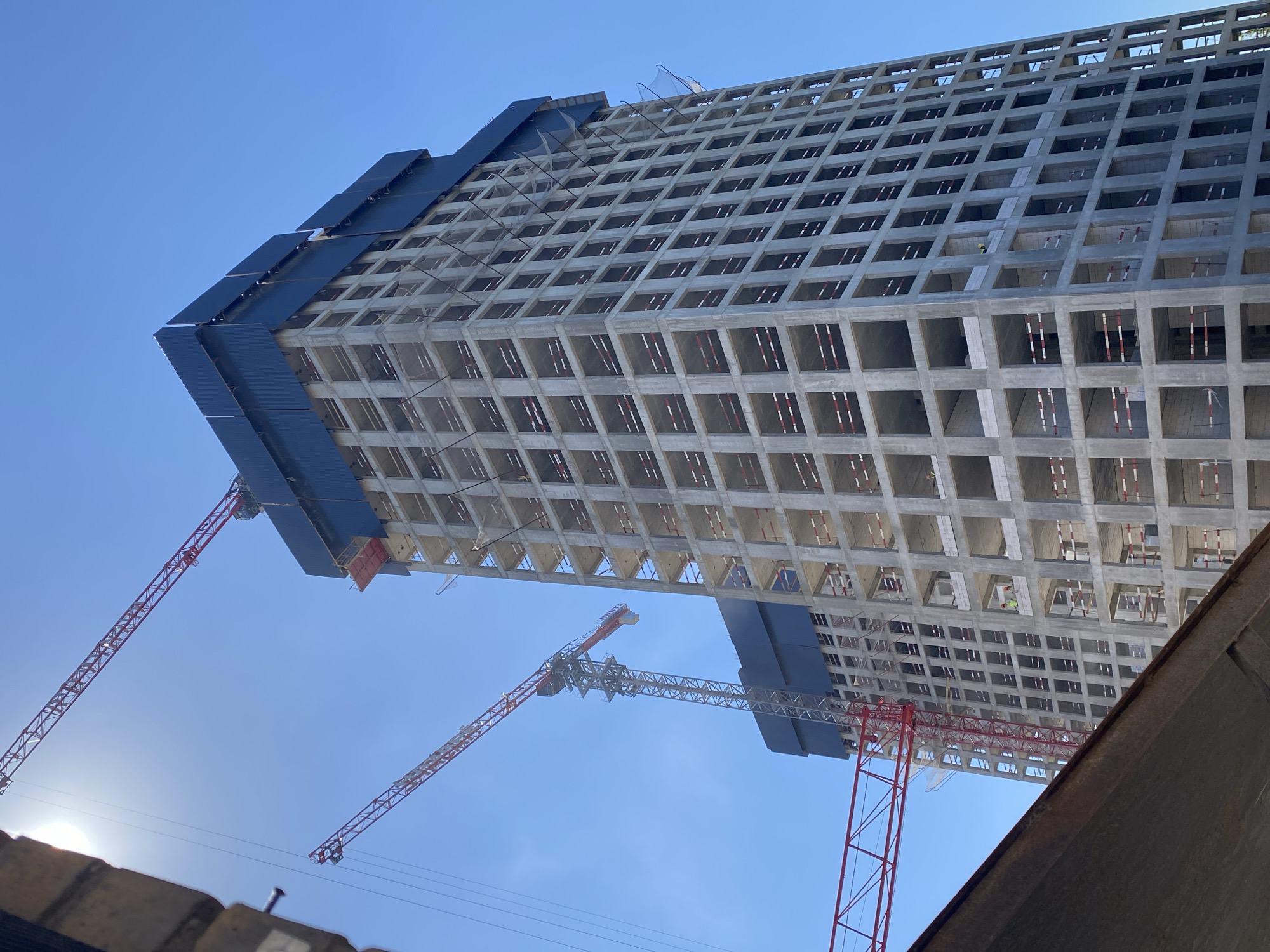 A high-angle, low-exposure, daytime, eye-level shot captures the unfinished concrete structure of a skyscraper under construction, located in Moscow, Russia. Two red tower cranes are visible against a clear blue sky. The main skyscraper, a tall, rectangular building, exhibits a grid-like facade with open concrete frames that will eventually become windows. Blue tarpaulin covers the top section of the skyscraper. Red and white safety netting is strung across some of the open window frames on the building's exterior. Two construction workers in hard hats and high-visibility vests are visible on a lower level of the skyscraper. The scene is framed by a dark, concrete structure in the foreground, partially obscuring the lower left corner of the image.