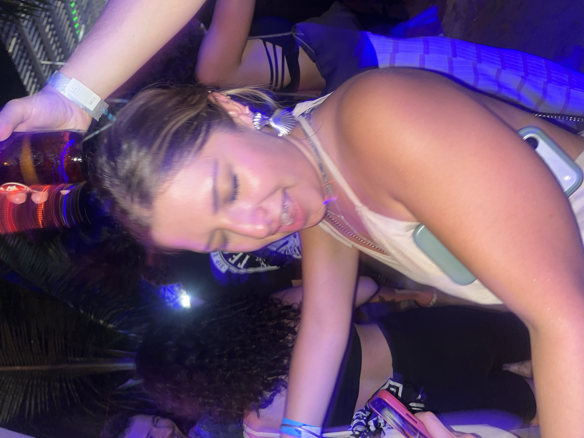 The image depicts a group of young adults in what appears to be a social setting, potentially a beach party or outdoor bar in Brisas de Zicatela, Mexico. The primary subject is a young woman with fair skin, tan, light brown hair, wearing a white tank top and silver necklace. She is smiling and appears to be interacting with the person holding a beer bottle over her head. This person is wearing a wristband, suggesting access to an event or venue. Other individuals are visible in the background, some lying down, and their attire suggests a relaxed, casual atmosphere. The lighting is a mix of artificial lights (possibly from the venue) and some natural light. The presence of what looks like palm leaves indicates a tropical environment. Several phones are present in the image.