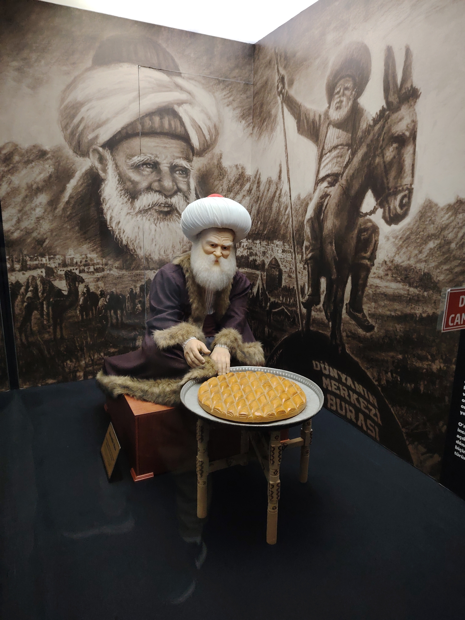 This image captures an indoor museum exhibit, likely in Eskisehir, Turkey, featuring a life-sized figure against a large wall mural. The central subject is a mannequin of an elderly man with a long, flowing white beard and mustache, wearing a prominent white turban with a red top and a traditional dark brown, fur-lined robe. He is seated on a wooden block, gazing thoughtfully at a round plate of diamond-patterned pastry, resembling baklava, which rests on a small, intricately carved wooden table in front of him.
The expansive, sepia-toned mural behind the figure depicts two main scenes: a large, detailed portrait of an old man with a white beard and turban on the left, closely resembling the mannequin; and on the right, a similar elderly man riding a donkey while holding a staff. The mural's background features mountains, a line of camels, and a distant settlement, evoking a historical and rural atmosphere.
Prominent text visible on the mural reads "DÜNYANIN MERKEZİ BURASI," which translates to "THE CENTER OF THE WORLD IS HERE," a quote famously attributed to Nasreddin Hodja. A partial red sign on the far right edge of the frame shows "Dİ..." and "CAN...". The dark, reflective floor completes the exhibit setting. The overall display pays homage to a significant historical or cultural figure, very likely Nasreddin Hodja, given the location context and common iconography.