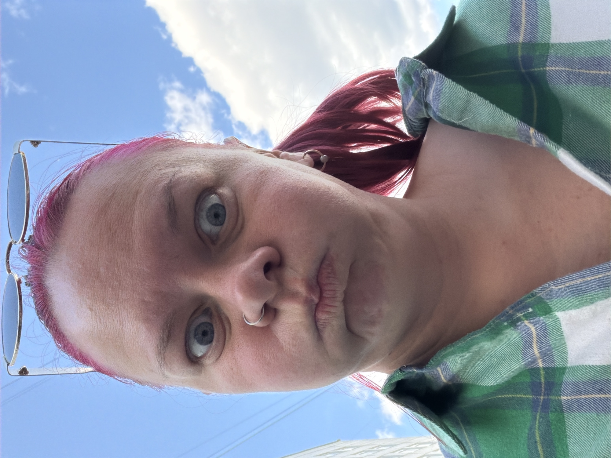 This is a low-angle, close-up selfie of an adult female outdoors in what is identified as Chaikovsky, Russia. The subject has bright pink hair, pulled back into a ponytail on the right side, with a pair of oval-lensed sunglasses resting on top of her head. Her blue eyes are wide open, looking directly into the camera, and her lips are pursed in a deliberate pout, creating an exaggerated, playful expression. She has a silver hoop nose ring in her left nostril and small gold-toned hoop earrings. She is wearing a green, white, blue, and yellow plaid collared shirt, visible from the shoulders down to the upper chest. The background predominantly features a clear bright blue sky, with scattered white clouds visible on the right side. On the far left, a portion of a light-colored building is partially visible, and thin power lines or cables stretch across the upper left corner of the sky.