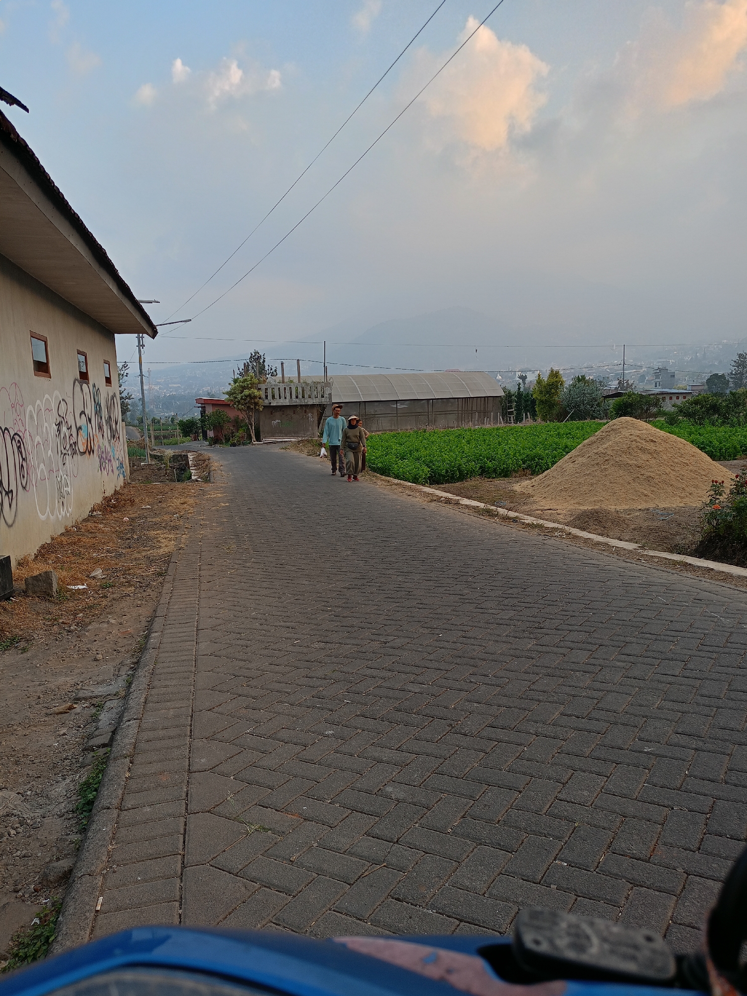 This is a full-shot image taken in the city of Batu, Indonesia, on a paved road that appears to be in a rural or suburban area. The sky is partly cloudy with a mix of blue and light-colored clouds. On the left side of the image, there is a light-colored building with several windows and graffiti on its wall. A paved sidewalk runs alongside the building. The road itself is paved with dark interlocking bricks arranged in a herringbone pattern. Two individuals are walking away from the viewer on the road. The person in front is wearing a light blue long-sleeved shirt and dark pants, and the person behind them is wearing a beige head covering, a dark long-sleeved top, and dark pants. To the right of the road, there is a large pile of light brown material, possibly agricultural produce or soil, situated next to a field of green crops. In the background, there are buildings, trees, and hills, suggesting a populated area with a natural landscape. The overall lighting suggests it is either early morning or late afternoon.