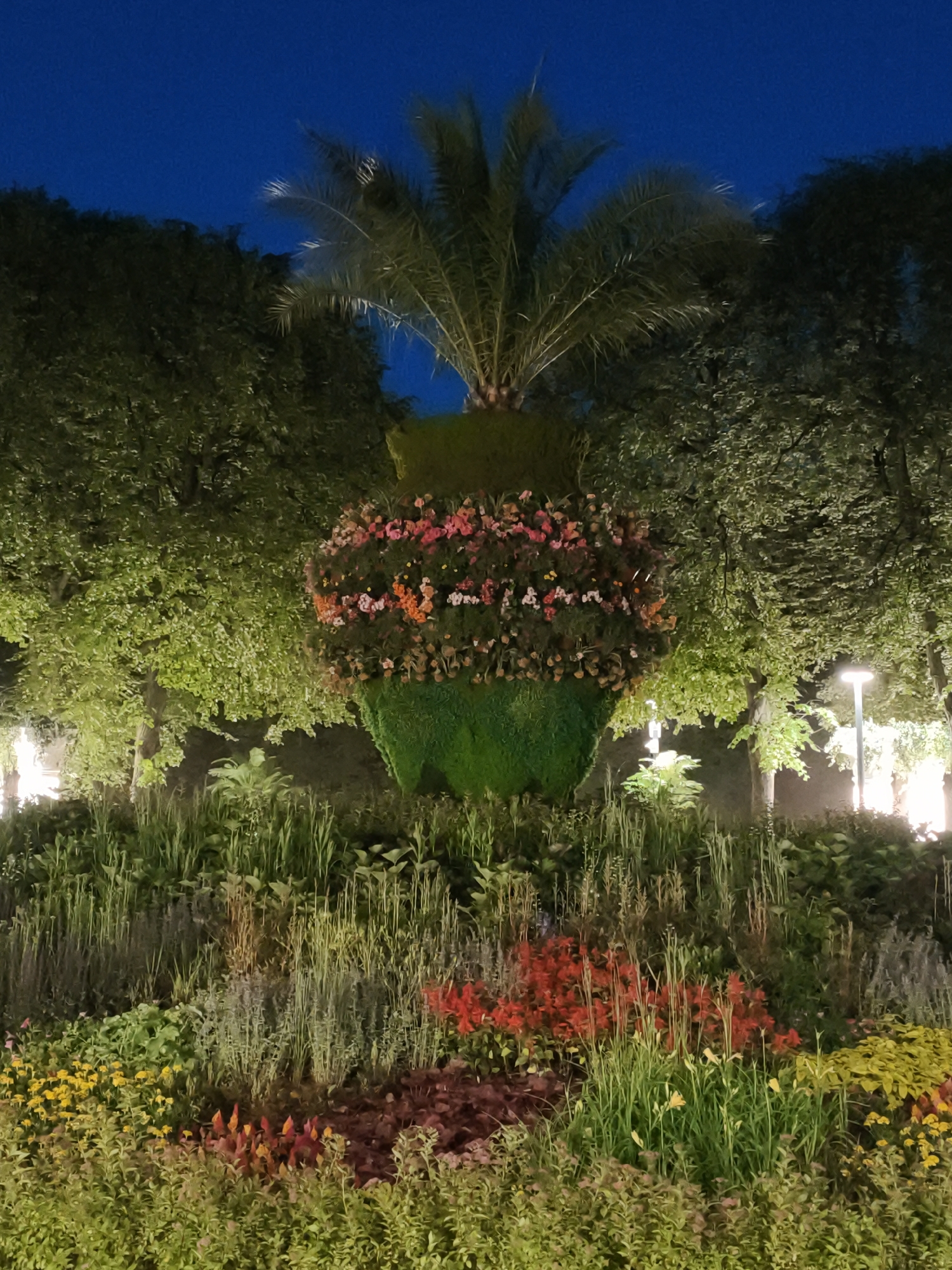 The image depicts an outdoor floral display at night. The centerpiece is a large, urn-shaped topiary covered in greenery, adorned with a vibrant band of pink, orange, and yellow flowers around its middle. A palm frond extends from the top of the topiary. Behind the display, lush green trees are illuminated, and lampposts with warm lighting are visible in the background. The foreground features a densely planted garden bed with various types of flowers and foliage in shades of green, red, yellow, and purple. The sky above is a deep, dark blue, indicating nighttime. There are no people or animals visible in the scene. The location is identified as Unknown, Unknown.