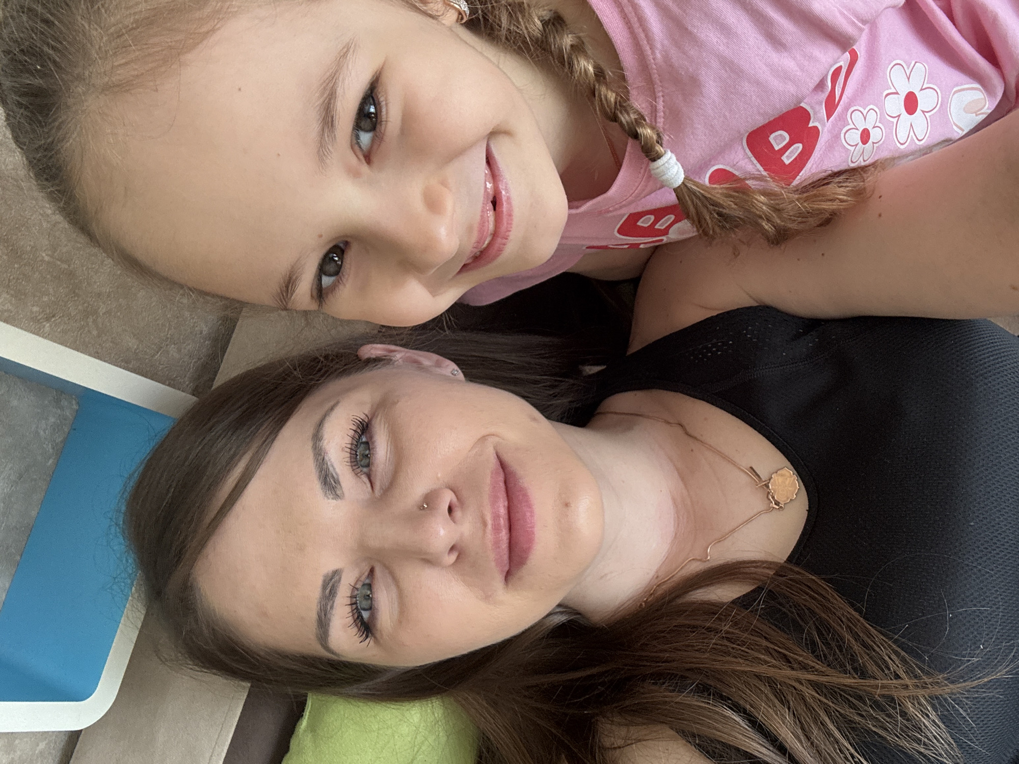 A photograph shows a young girl and a woman lying close together, presumably for a selfie or photo. The girl has light brown hair styled in a braid with a white tie, wearing a pink t-shirt with flower designs. The woman has long brown hair, wearing a black shirt and a necklace. The location appears to be indoors, on or near a couch with a green pillow visible. It is not possible to confirm the location as Раздолье, Russia based on the image alone.
