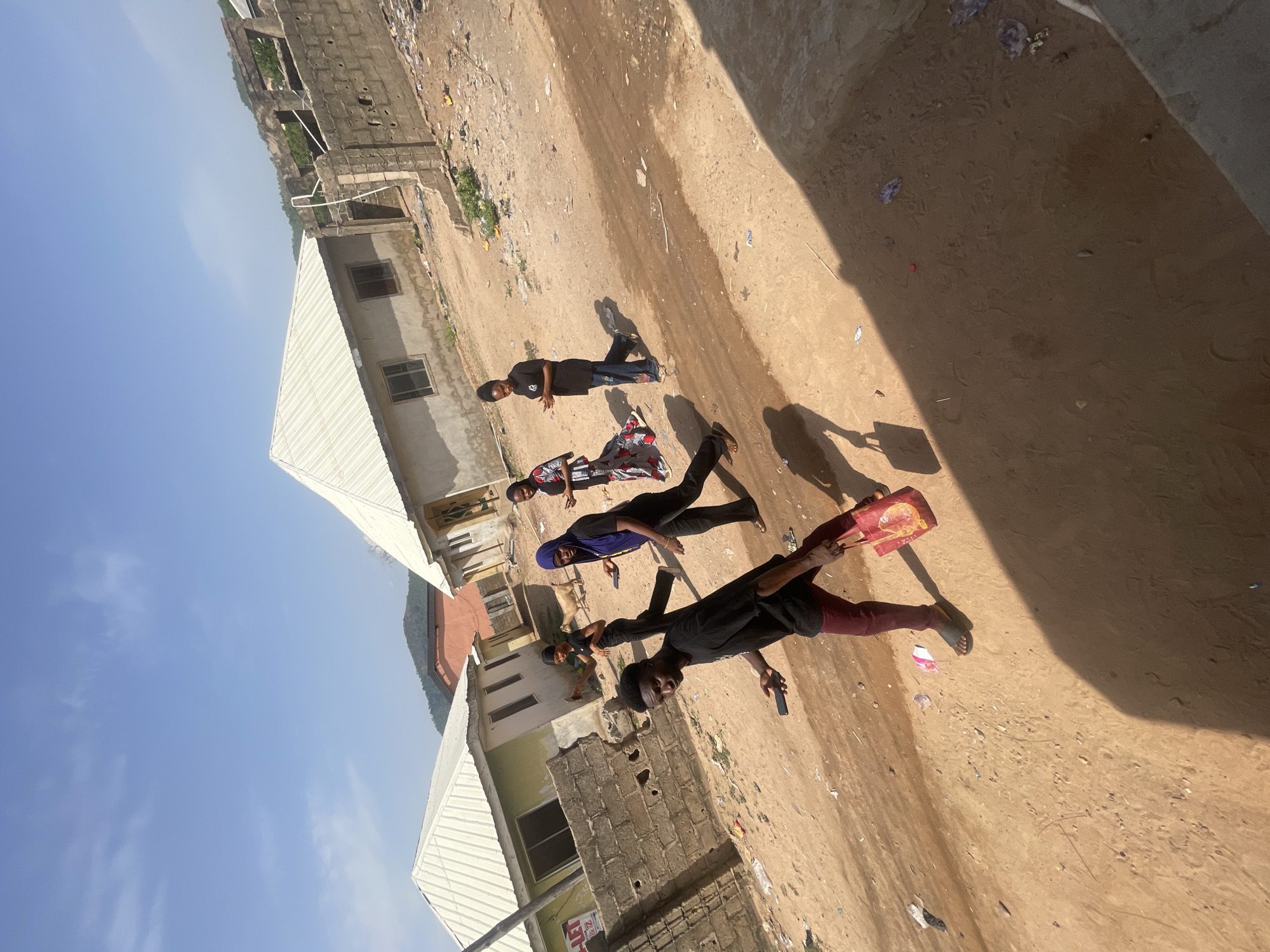 The image shows a group of four people standing on a dirt road in Lokoja, Nigeria. The people are dressed in casual clothing. A man in the foreground holds a red, rectangular object. Two buildings are visible on the left side of the road. The sky is blue and clear. There is trash scattered on the ground.