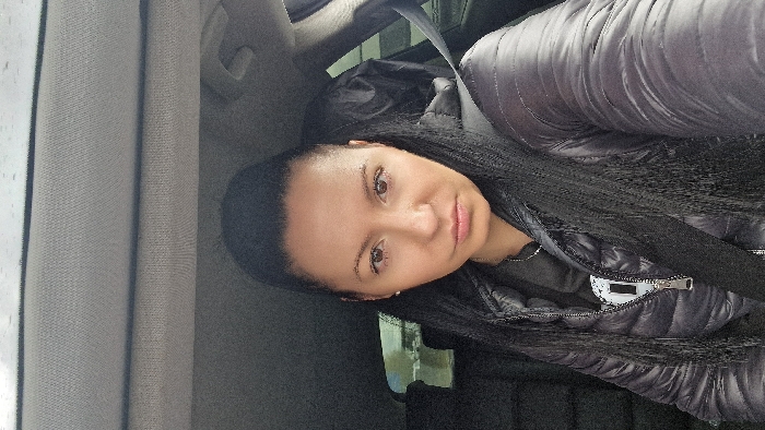 A female individual is depicted in a close-up, front-facing perspective, seated within the interior of a vehicle, appearing to be a car. The specific location within Bègles, France, is not visually identifiable from the image content.
**Subject:** The individual is a female with long, straight black hair pulled back into a high ponytail, with some strands falling over her shoulders and chest. She has a light to medium skin tone, brown eyes, and a neutral facial expression, looking directly at the camera. She is wearing a dark, possibly black, puffer-style jacket that is partially zipped, revealing a dark hooded garment underneath with a white graphic design on the left side of the chest. A thin, silver-colored chain necklace is visible around her neck. A dark seatbelt crosses diagonally from the upper right to the lower left of the frame, positioned across her chest and right shoulder.
**Environment:** The setting is the interior of a car. The upper portion of the frame shows a light gray, textured car ceiling lining. To the left, through what appears to be a rear passenger window, a blurred outdoor scene is visible, consisting of indistinct light and dark shapes. To the right, within the car's interior, a crumpled dark object, possibly a garment or bag, is visible on the rear seat. The immediate surroundings within the vehicle appear dark-toned, likely black or dark gray.