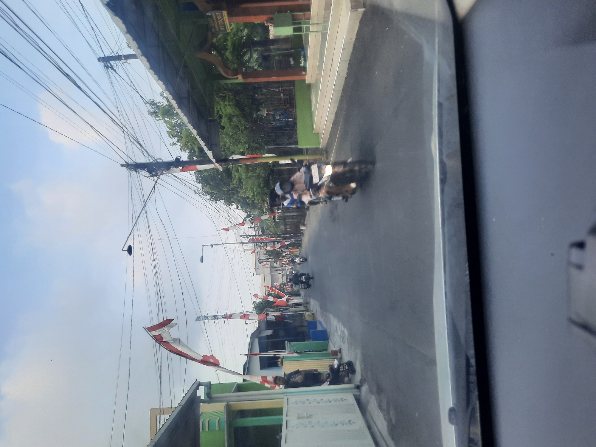 This image captures a street scene in Sidokepung, Indonesia, likely during a period of national celebration, as evidenced by the numerous red and white Indonesian flags adorning the street. The view is from inside a vehicle, looking out through the windshield. The street is paved and appears to be a residential or commercial area, with buildings lining both sides. Several motorcycles are present, with individuals riding them, suggesting typical daily activity.

On the left side of the street, a green and white building is visible, featuring a metal fence and a gate. A man stands near the gate, partially obscured. Further down the street, more flags are displayed, some hanging vertically and others draped across poles.

On the right side of the street, a building with a green roof supported by dark brown pillars stands out. This structure appears to be a place of worship or a community gathering space, with the word "AT-TAQWA" visible on a sign. A set of stairs leads up to this building, and some green chairs are positioned outside.

The sky above is partly cloudy with a bright, overcast feel. Overhead, a complex network of electrical wires crisscrosses the scene, supported by utility poles. The overall impression is one of a lively local street with decorations and ongoing daily life.