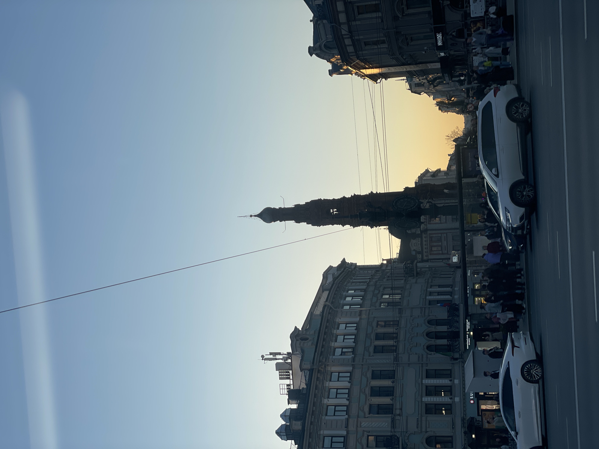 The image captures a street scene in Kazan, Russia, at dusk. The sky is a gradient from light blue to orange near the horizon. In the middle of the frame stands a tall, ornate tower with a spire. Buildings with architectural details line both sides of the street. Wires are strung across the sky. Pedestrians populate the sidewalks and areas along the buildings. Two white cars are parked on the right side of the street. The overall scene suggests a blend of historical architecture and modern urban life in Kazan.